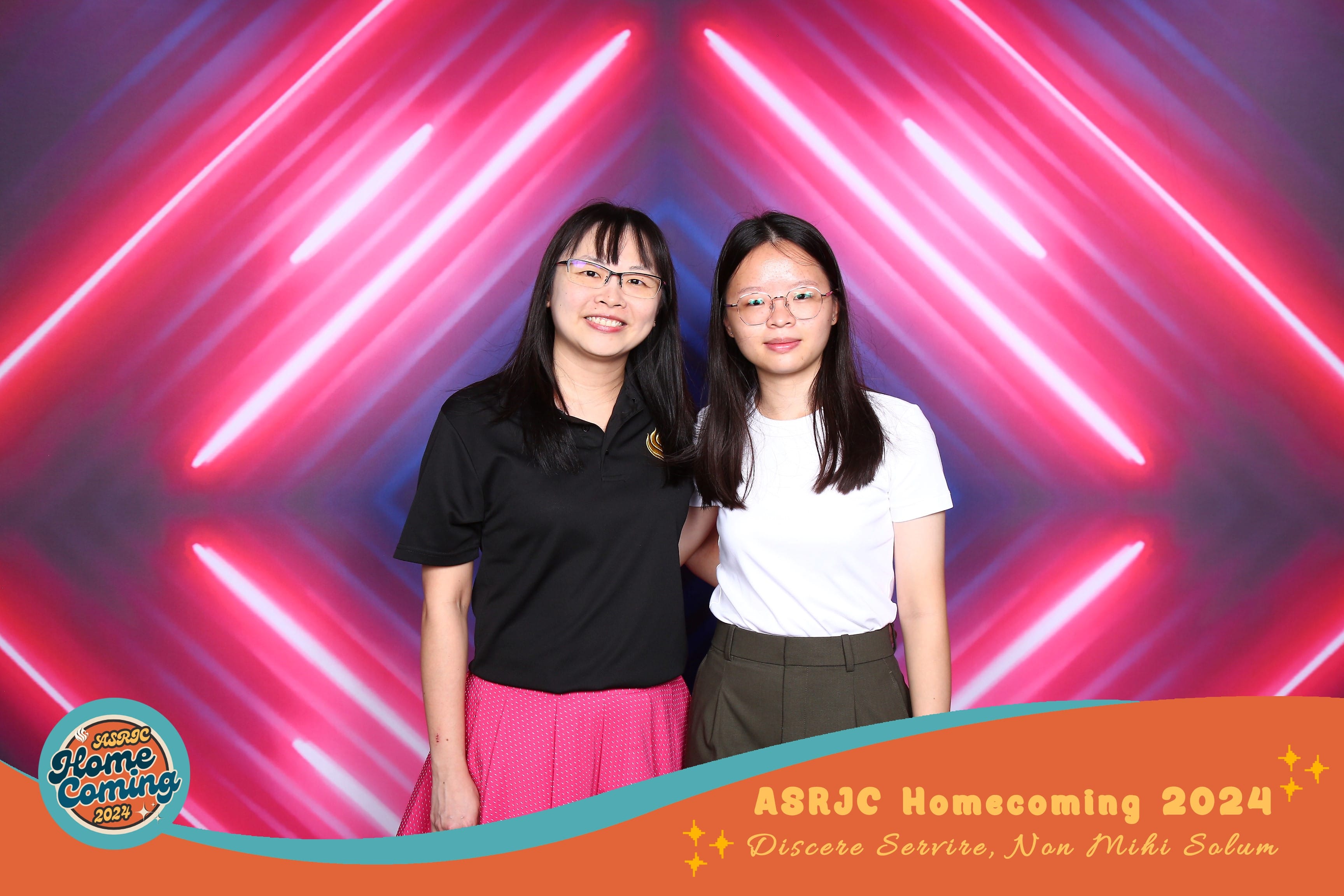 Two smiling women with glasses pose before a neon pink light backdrop at ASRJC Homecoming 2024.