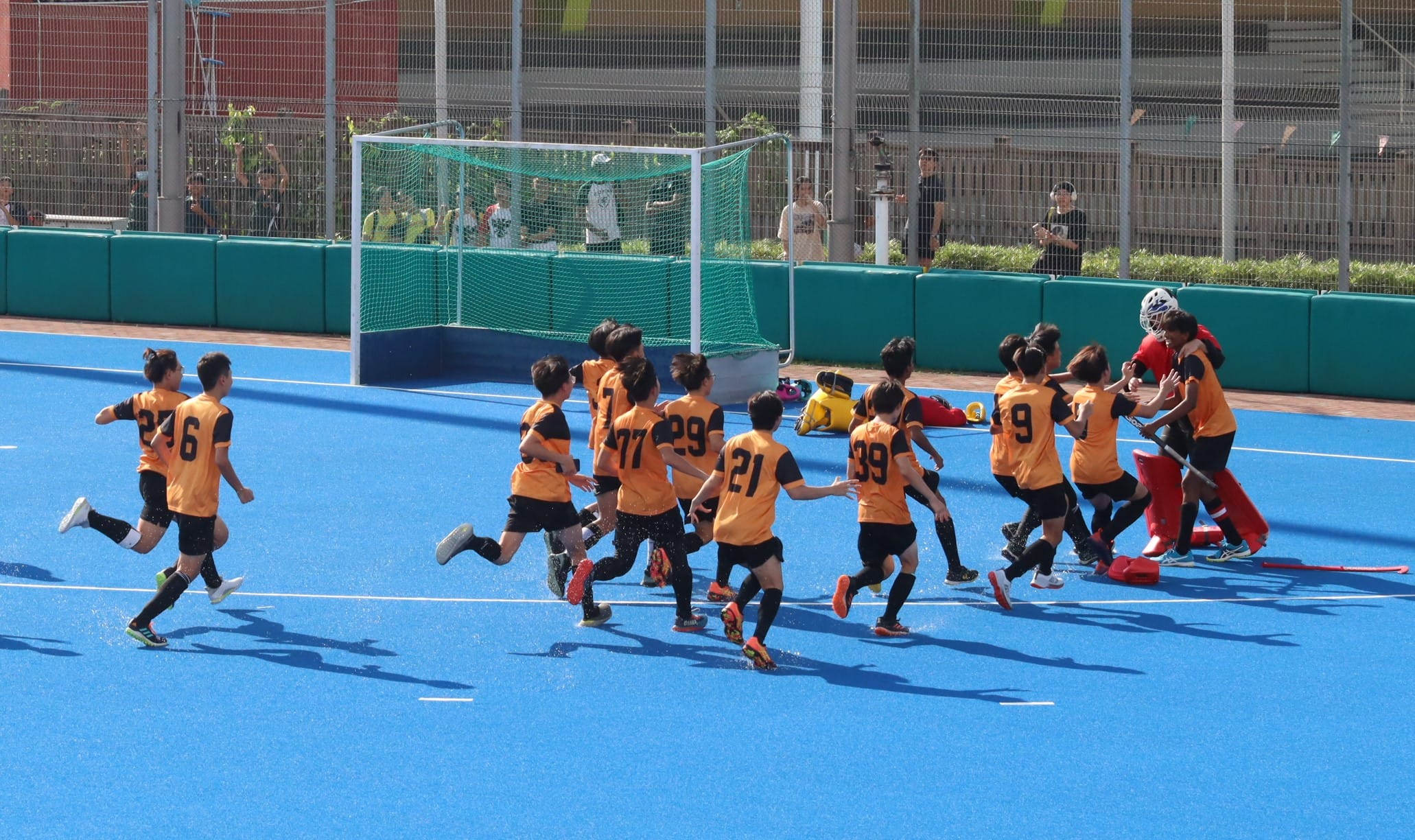 Young players in orange and black jerseys run and celebrate on a blue field hockey pitch near a goal.
