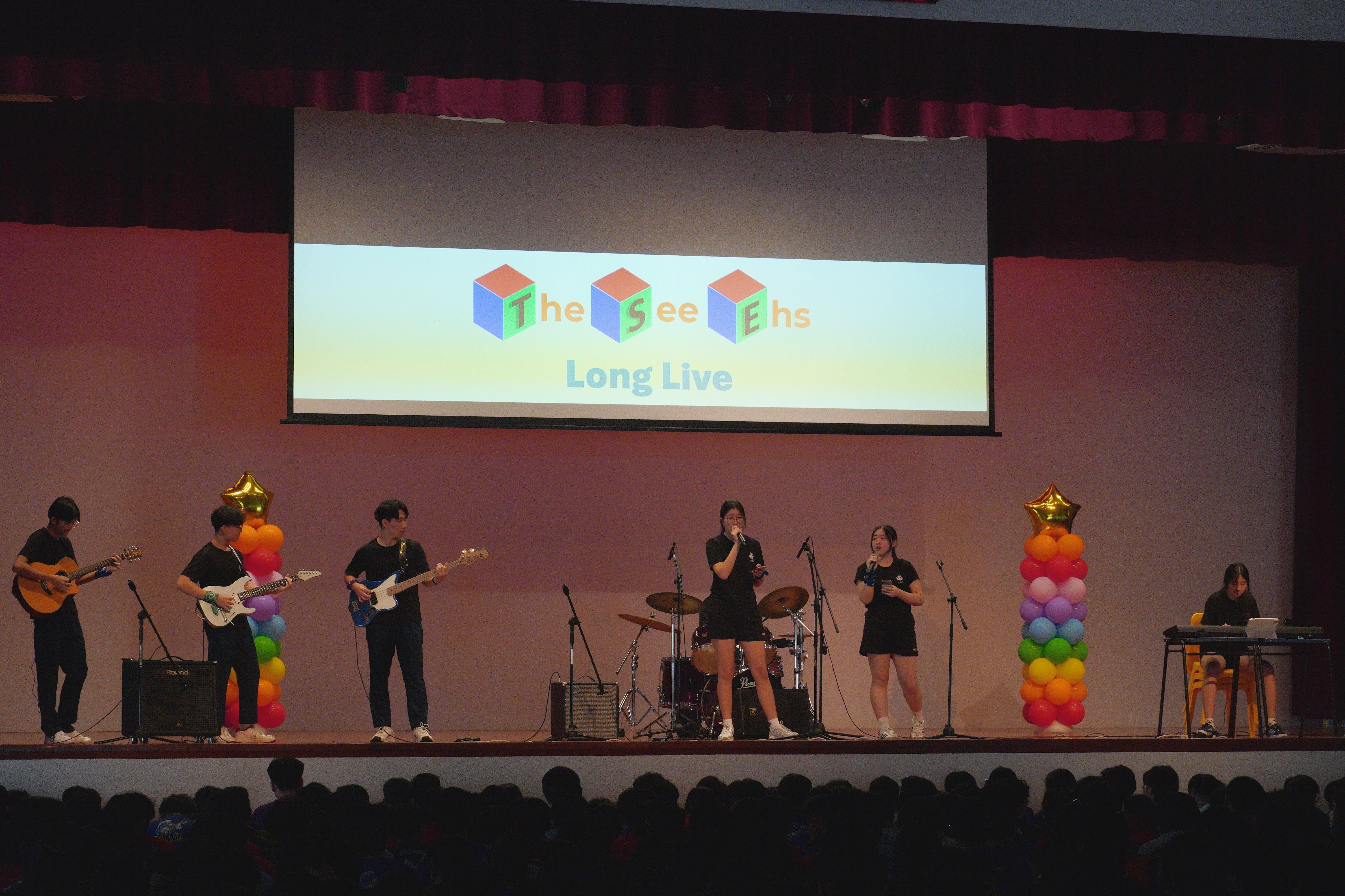 Student band performs on stage with guitars, drums, and vocals under a screen reading "The See Ehs" and "Long Live."