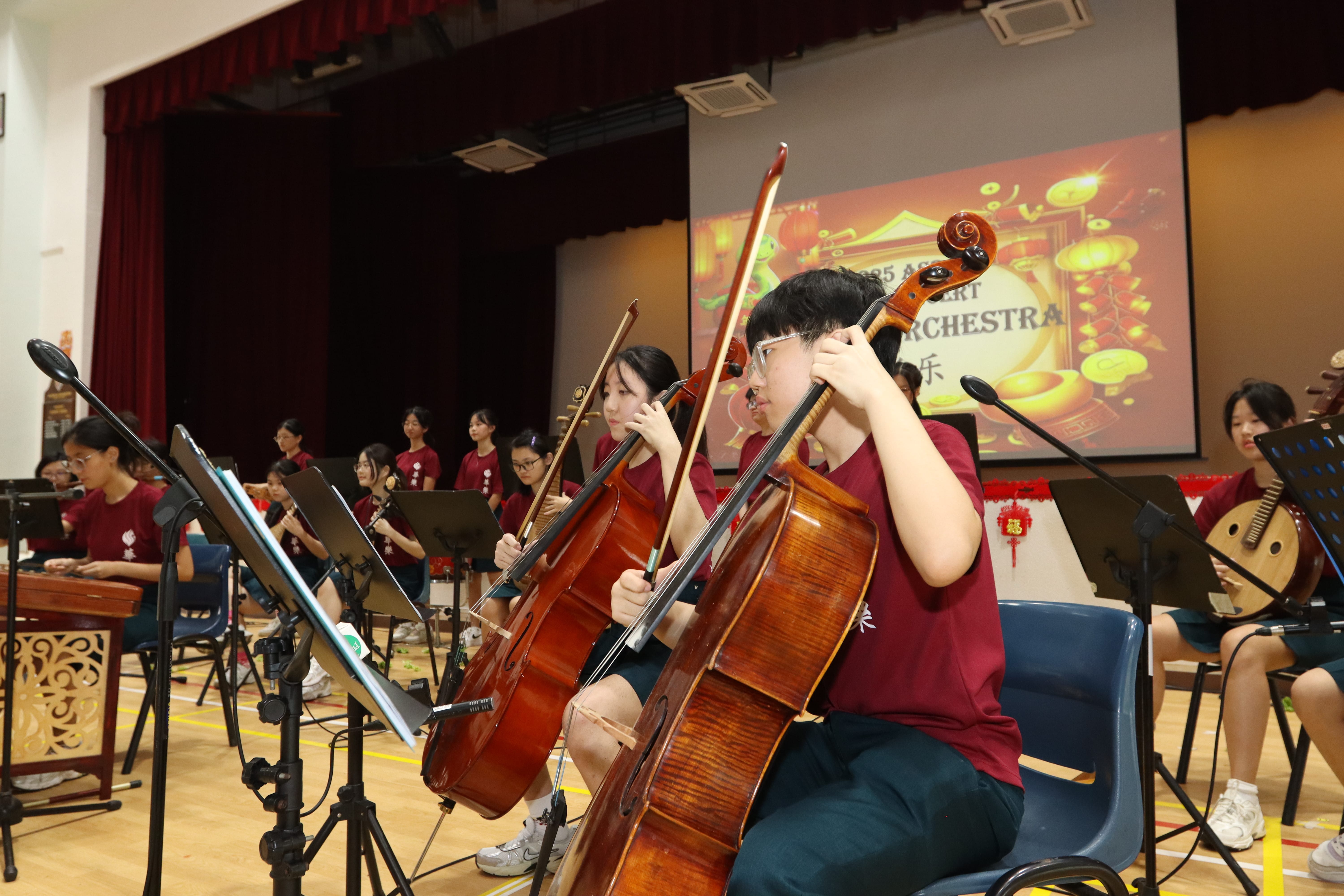 Students in maroon shirts perform on stage, with two cellists in front and music stands and screen behind.