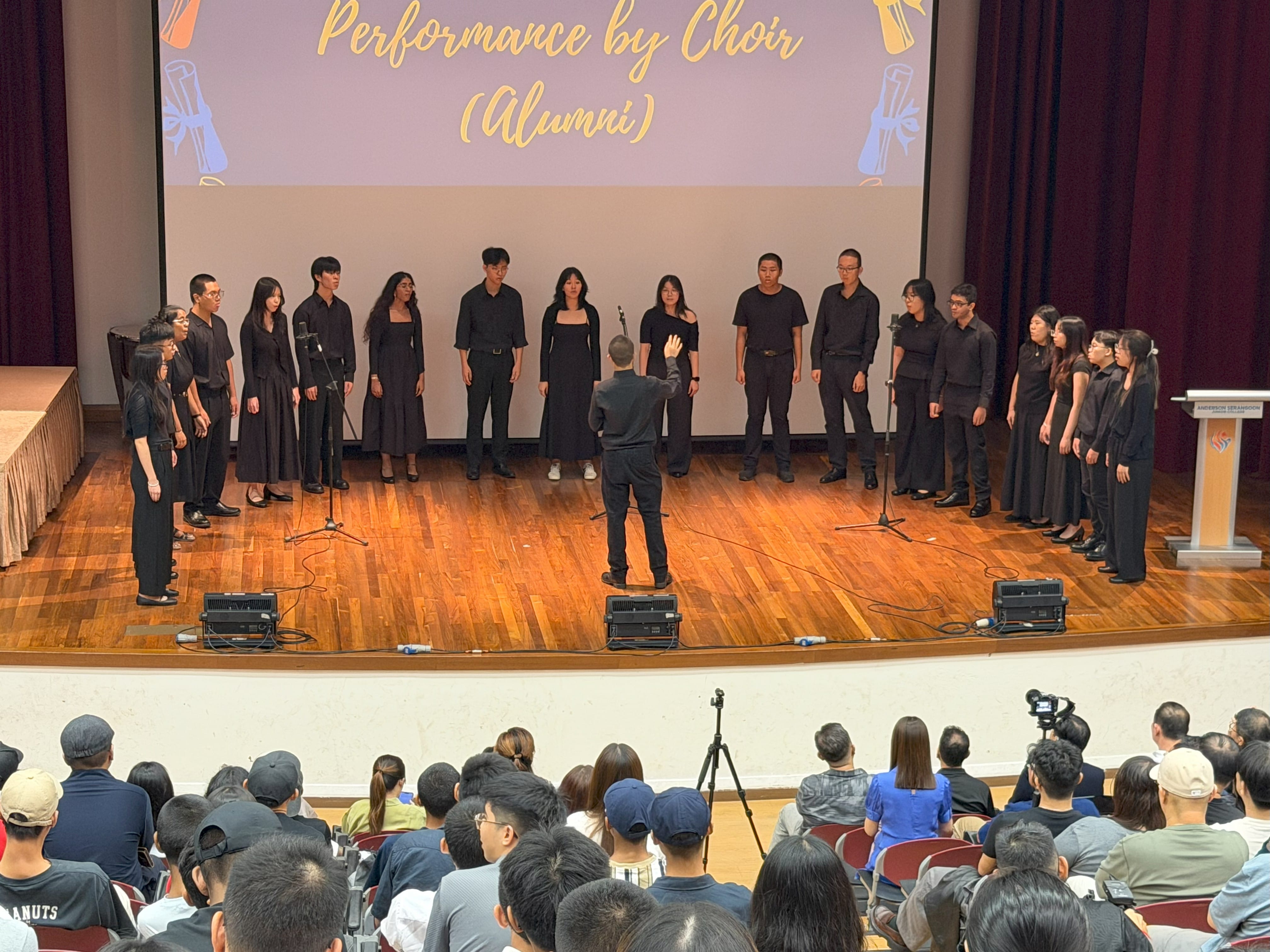 A choir dressed in black performs on stage, standing in a semi-circle facing the audience.