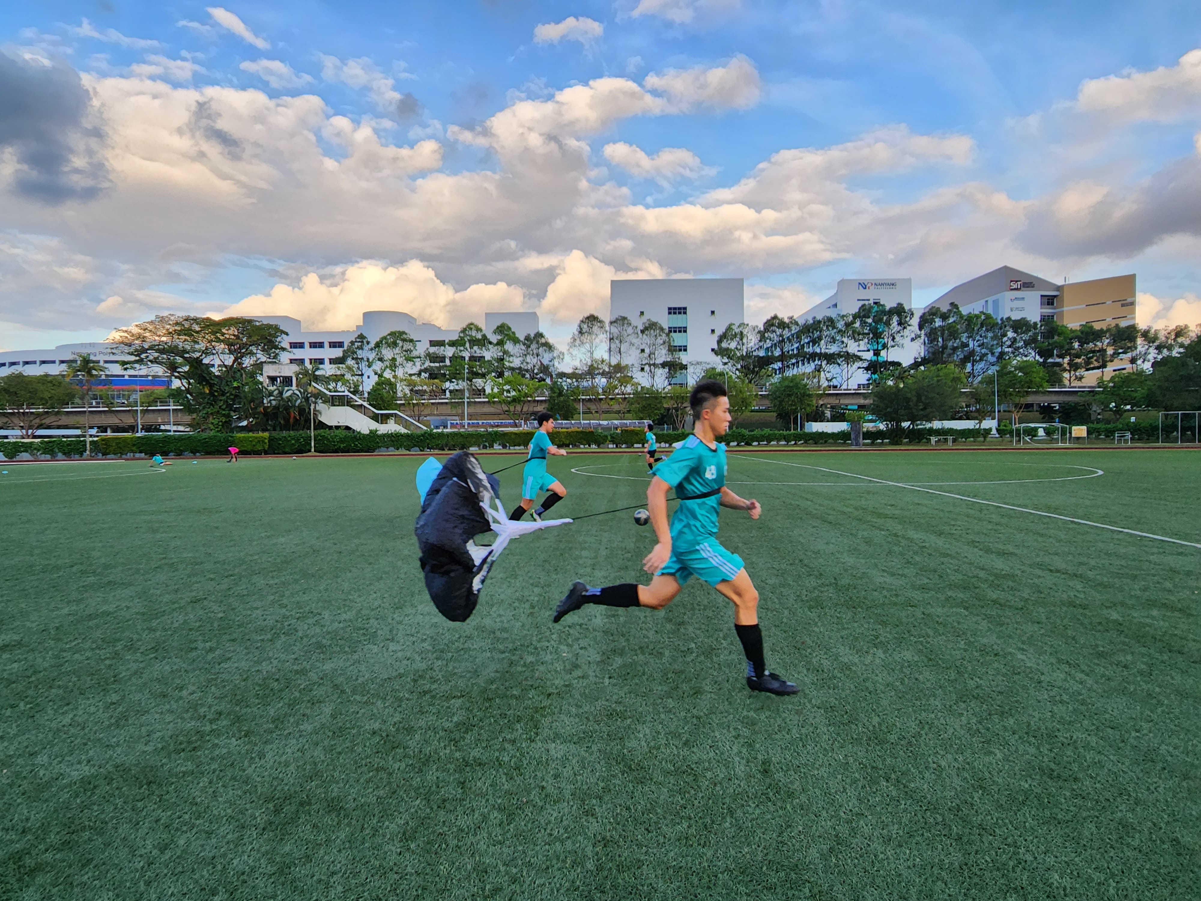 Two soccer players in teal uniforms run on a green field, dragging resistance parachutes during training.