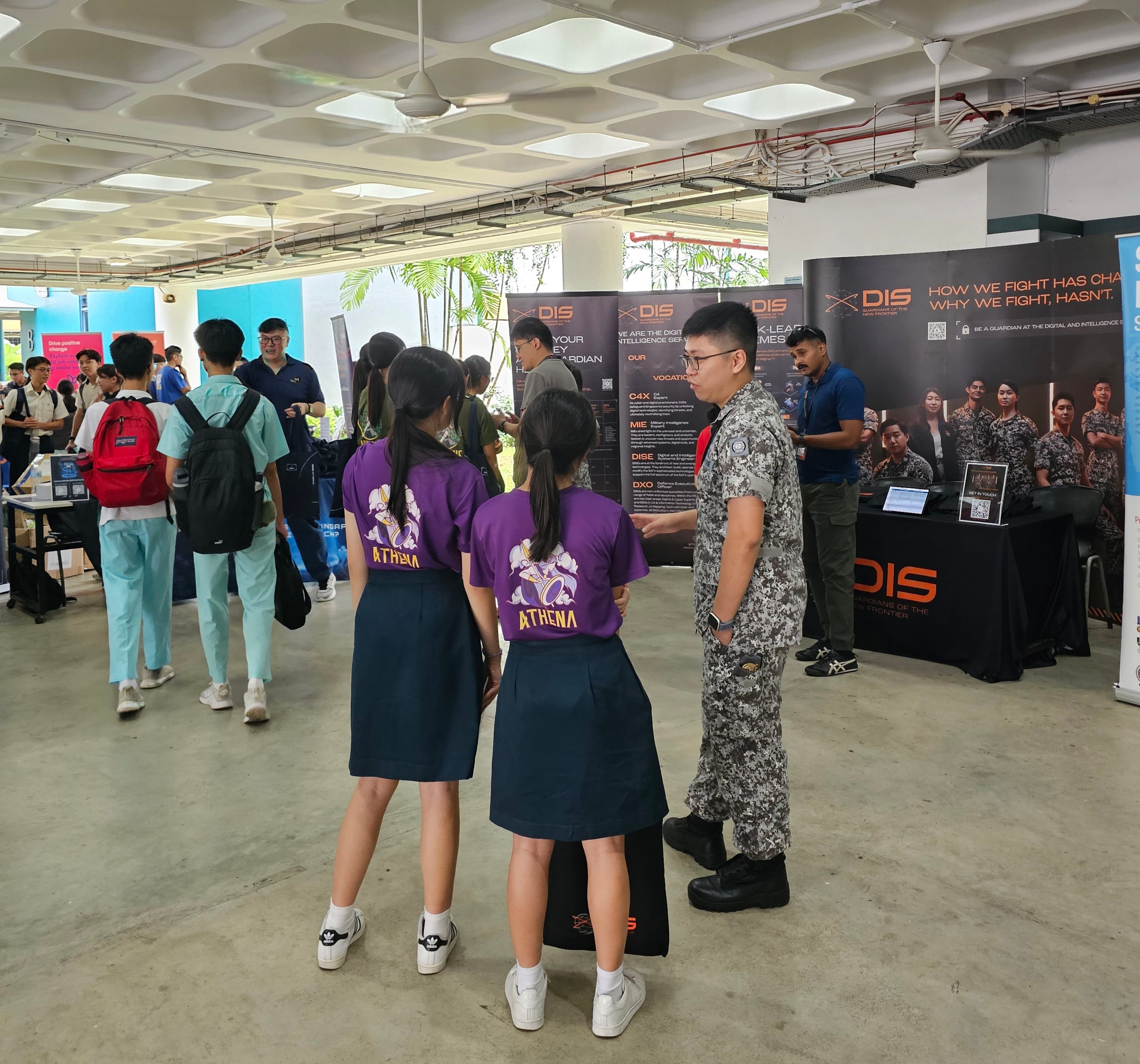 Two students in purple Athena shirts talk to a uniformed soldier at a DIS recruitment booth indoors.