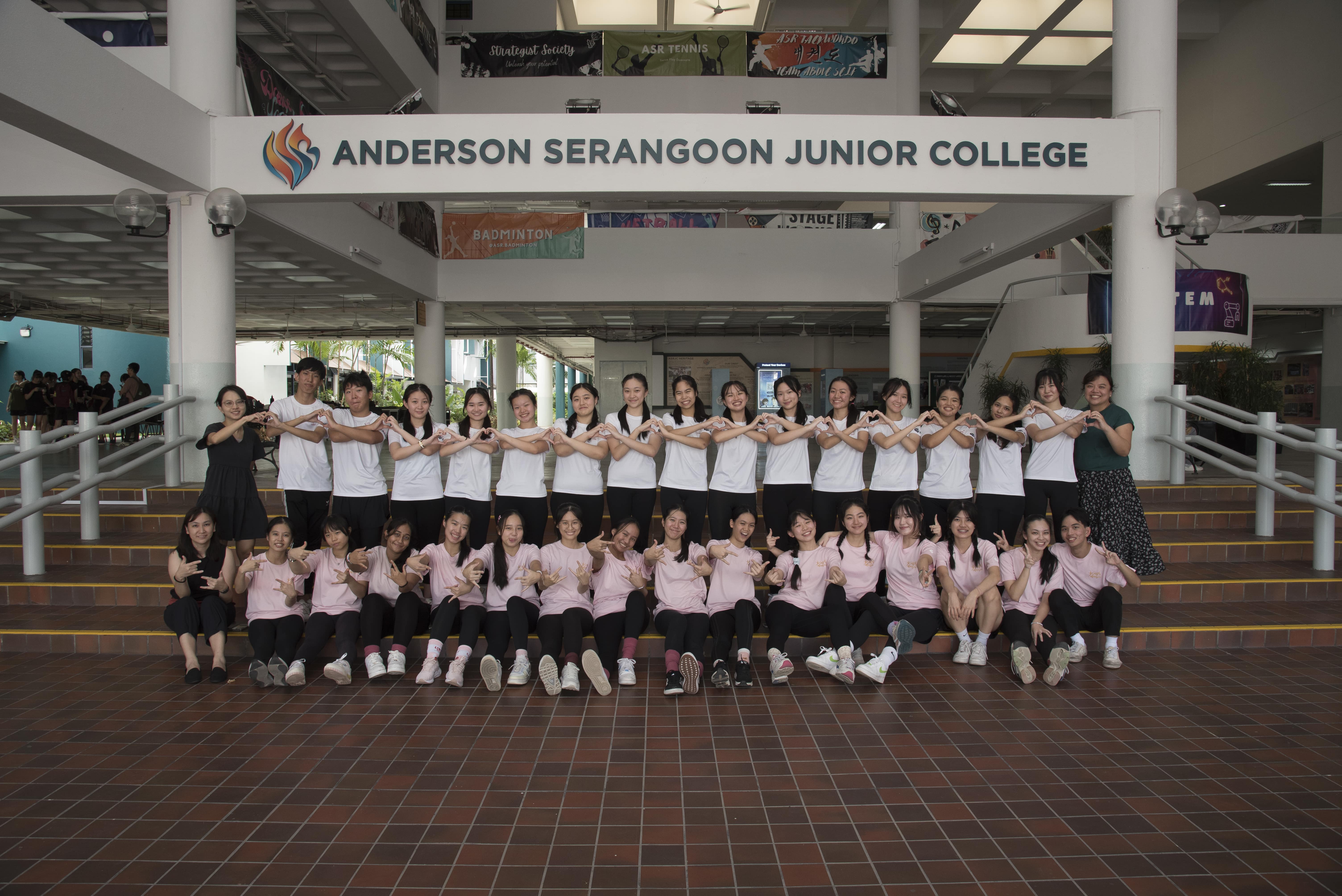 A large group of students pose making heart gestures under the Anderson Serangoon Junior College sign.