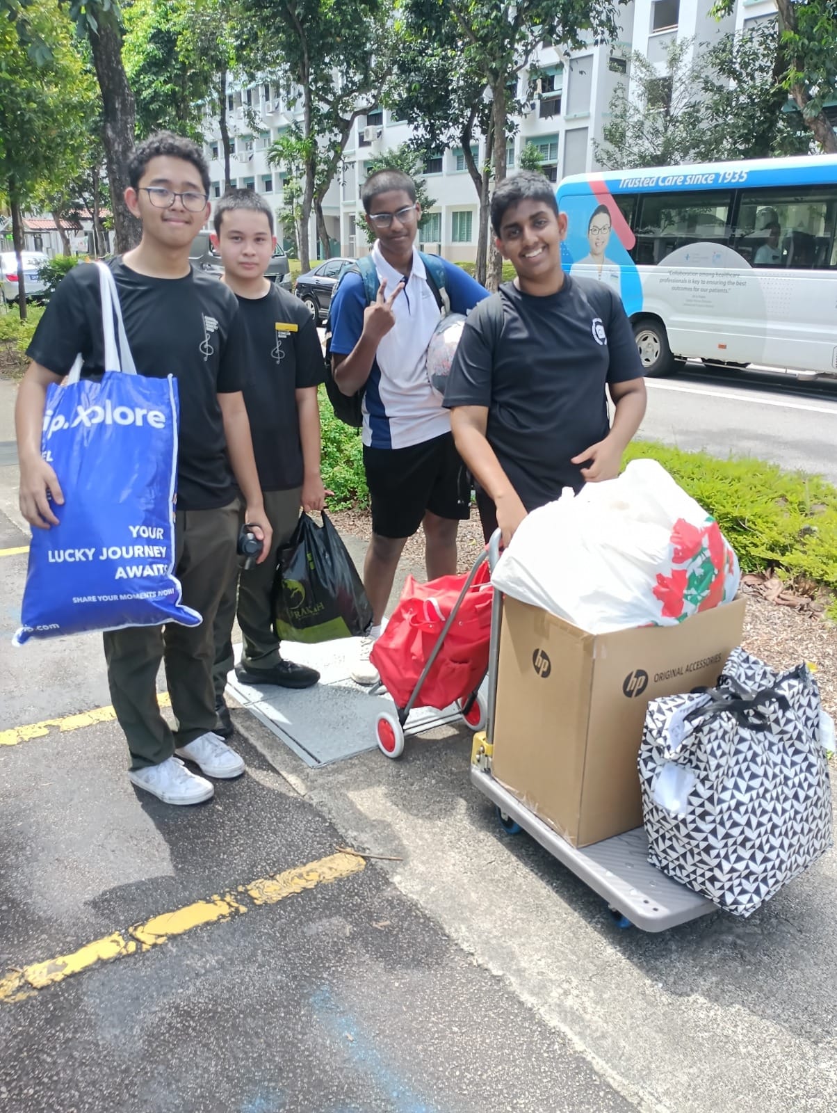 Four teenage boys stand outdoors near a trolley loaded with boxes and bags, with trees and a van behind them.