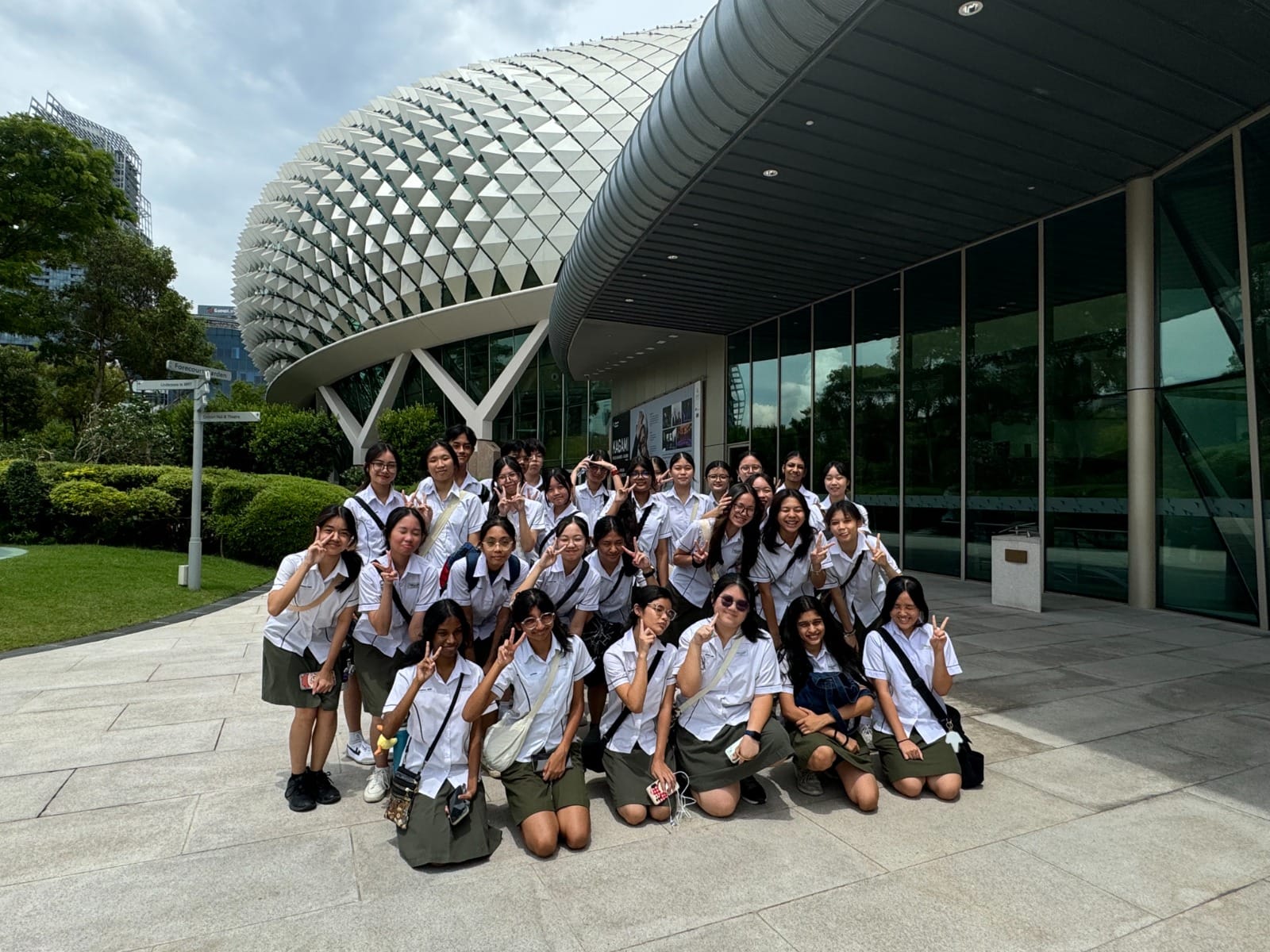 A large group of students in white uniforms pose with peace signs outside the Esplanade building in Singapore.