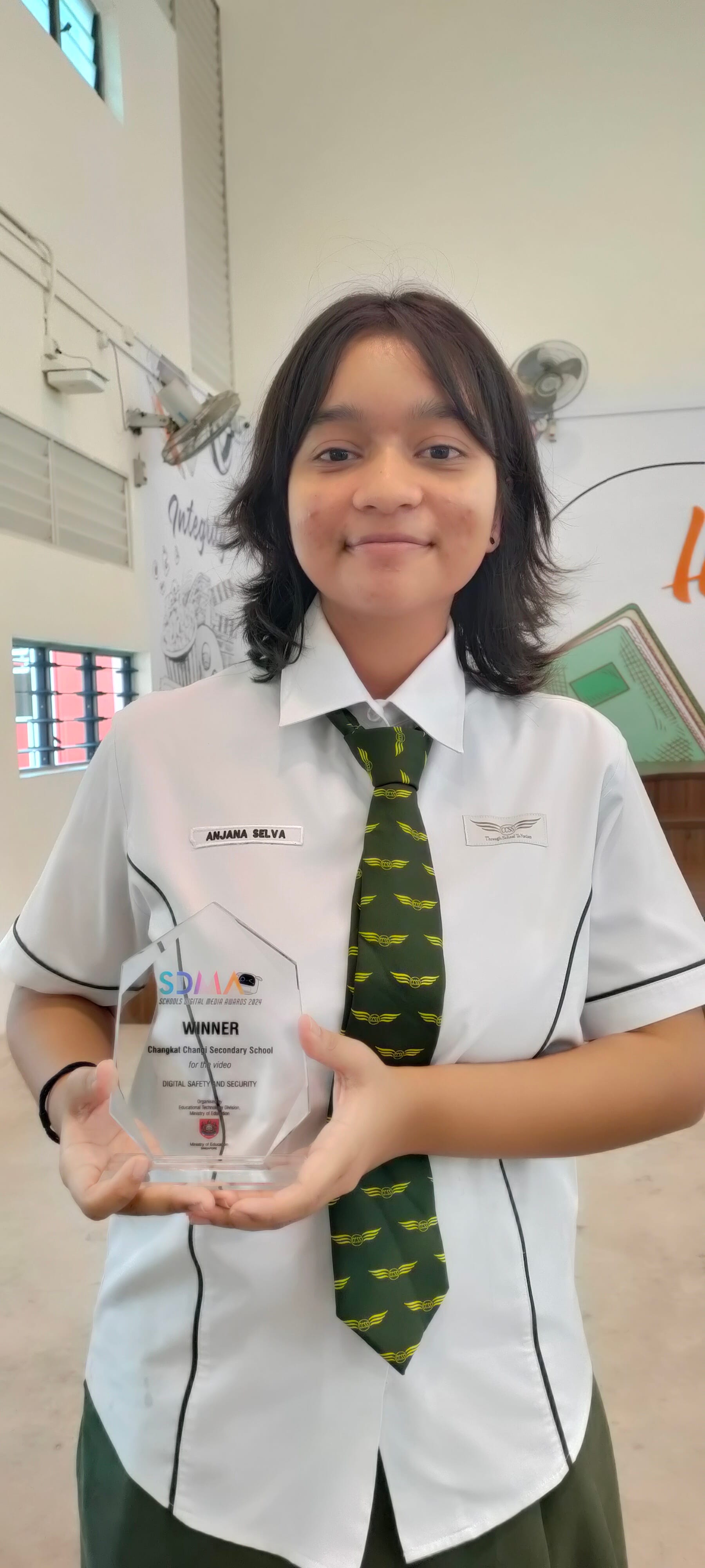 A young student in school uniform holds a clear acrylic SDMA Winner trophy inside a school hall.