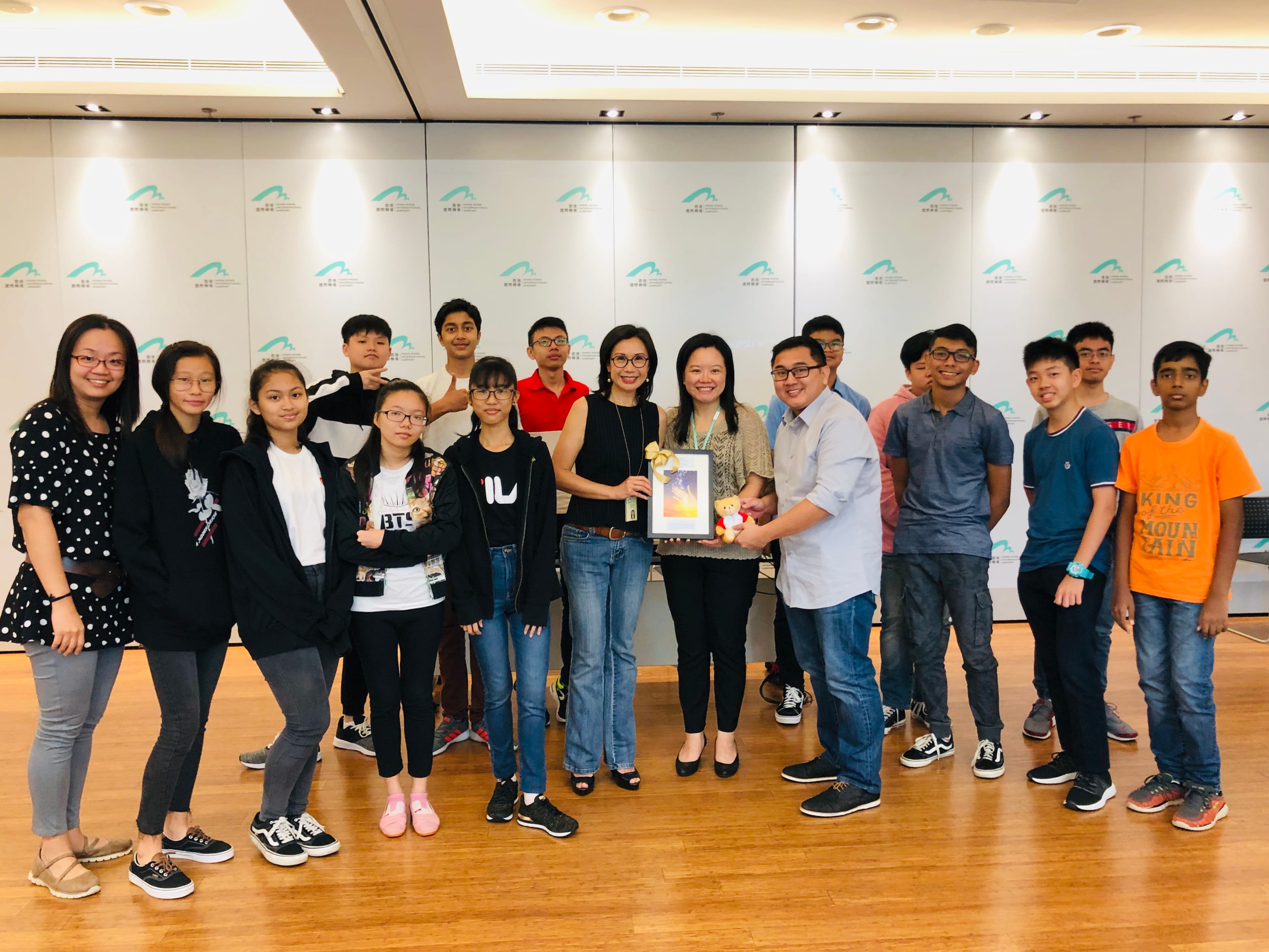 A group of teenagers and two adult women pose together indoors, with two people holding a framed picture and a small toy.