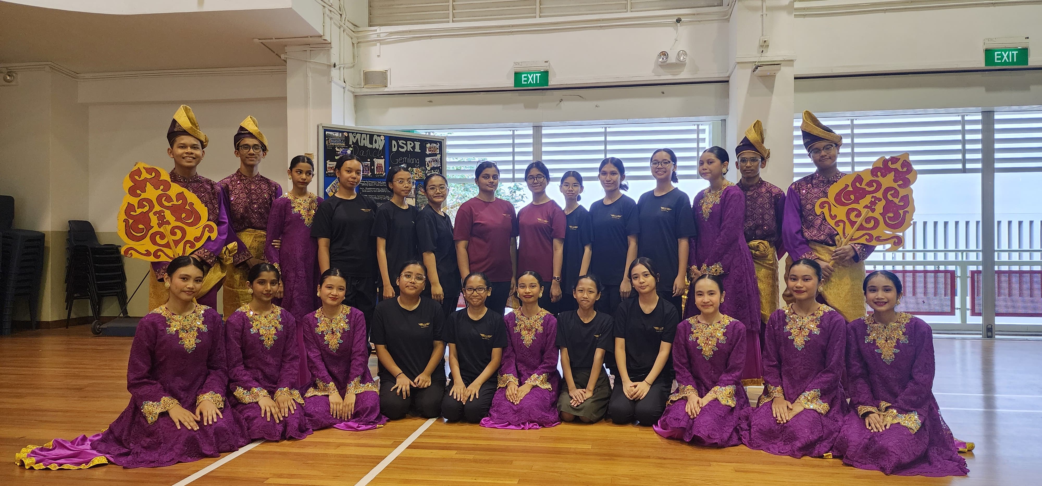 A large group of students in purple and gold traditional Malay costumes pose together in an indoor hall.