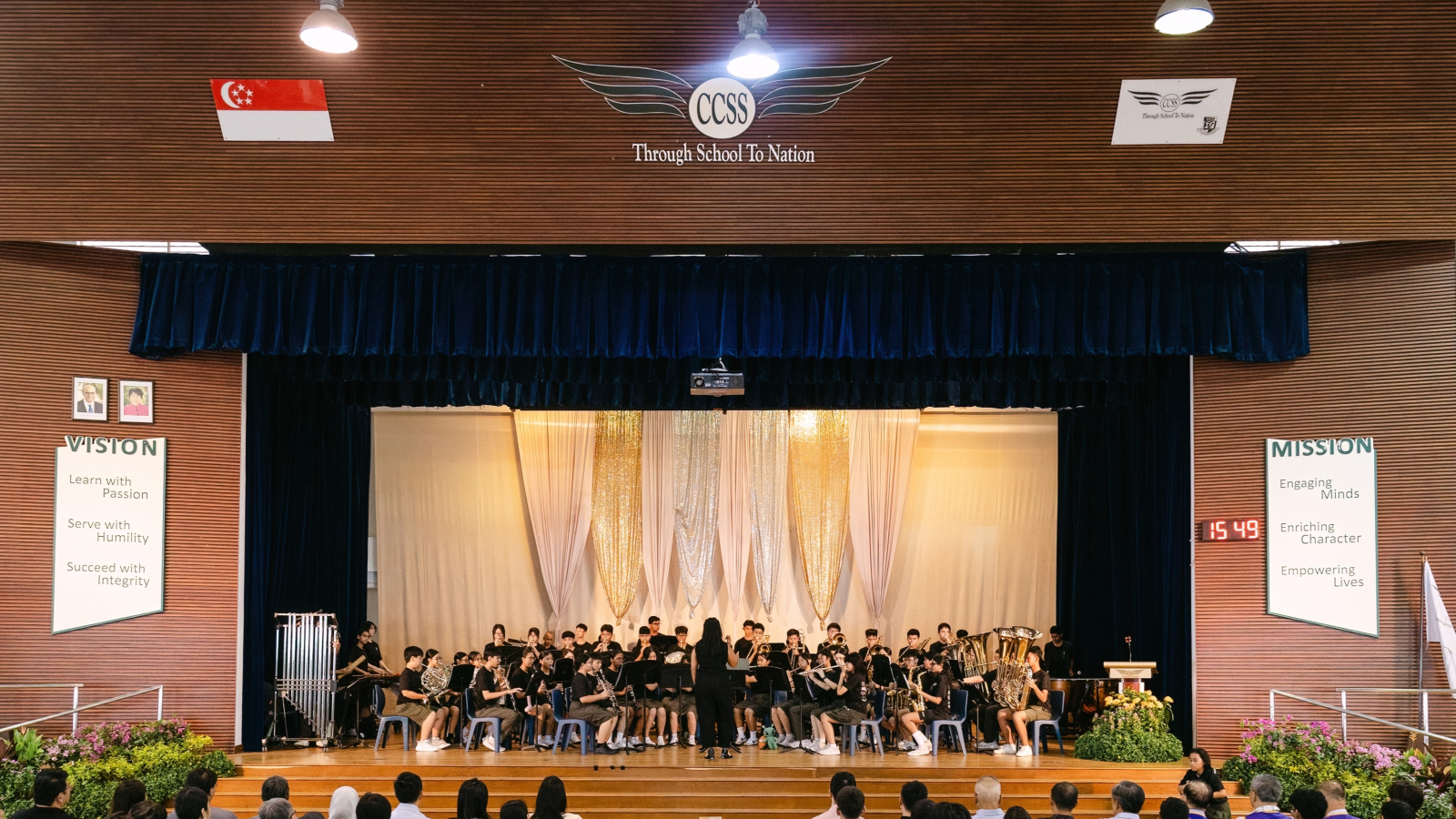 A school band in black uniforms performs on a decorated stage under the CCSS school motto banner.