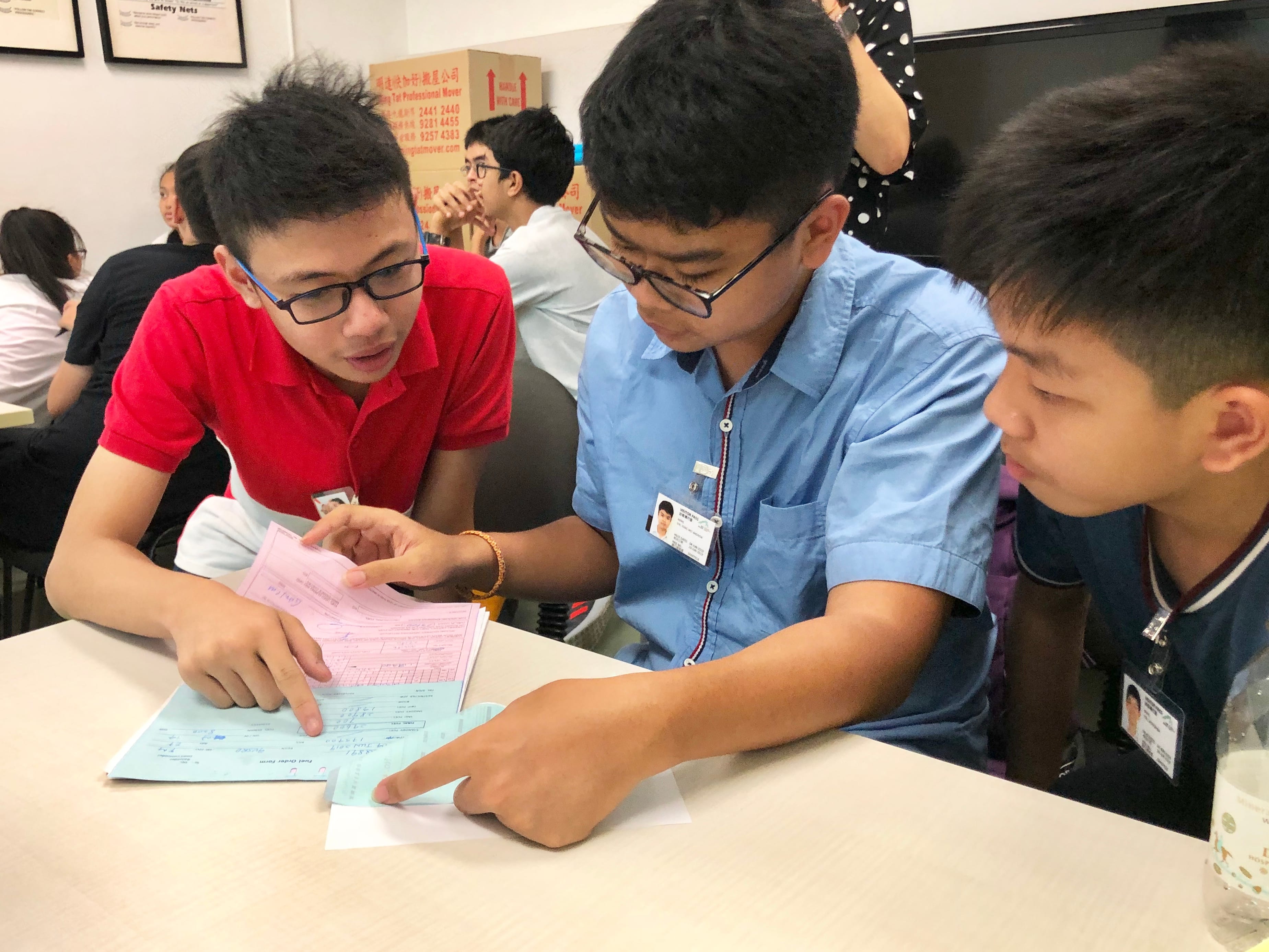 Three teenage boys with name badges review printed forms together at a table in a busy room.