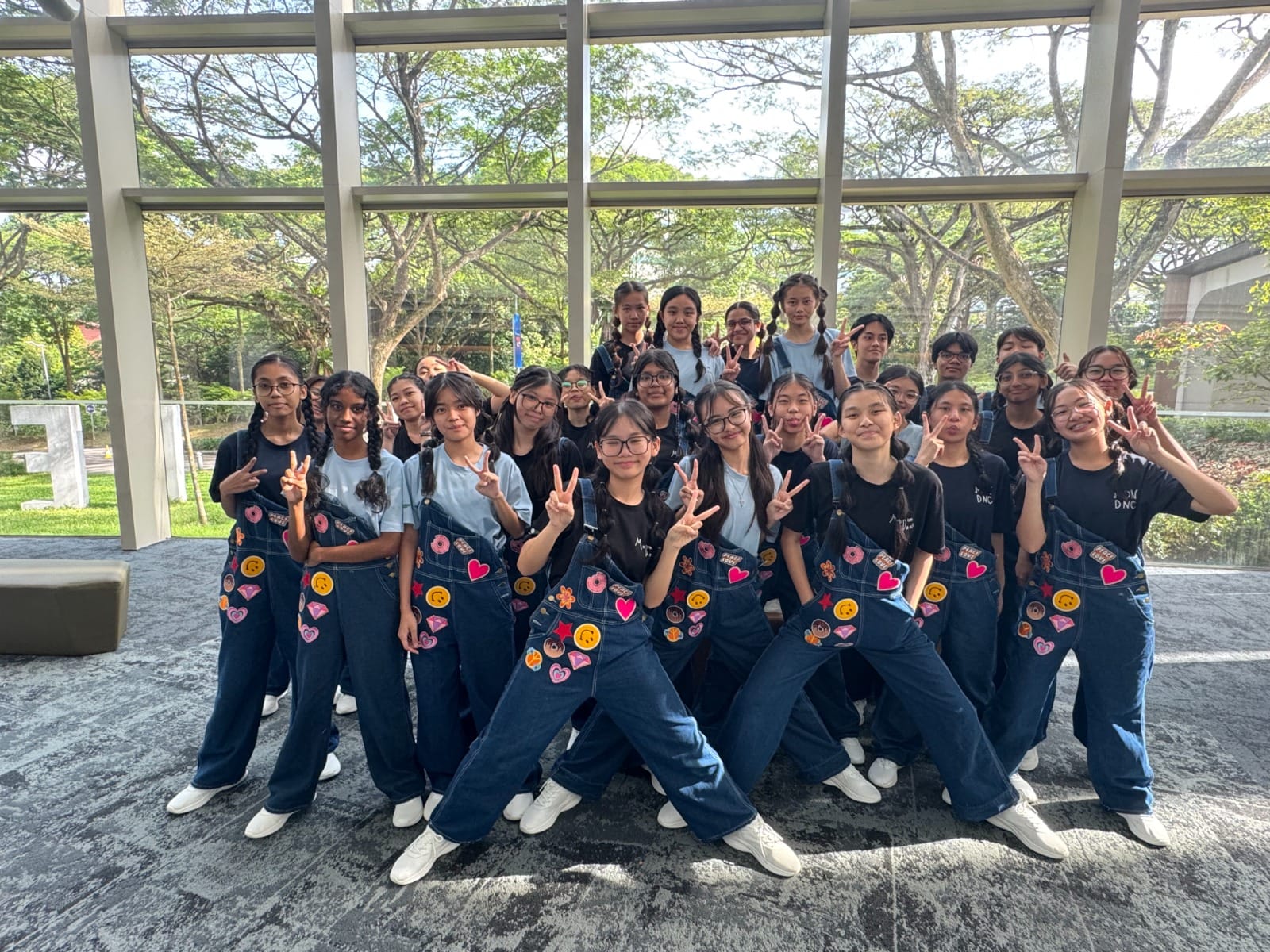 A large group of students in denim overalls with colorful patches pose with peace signs indoors near a glass wall.