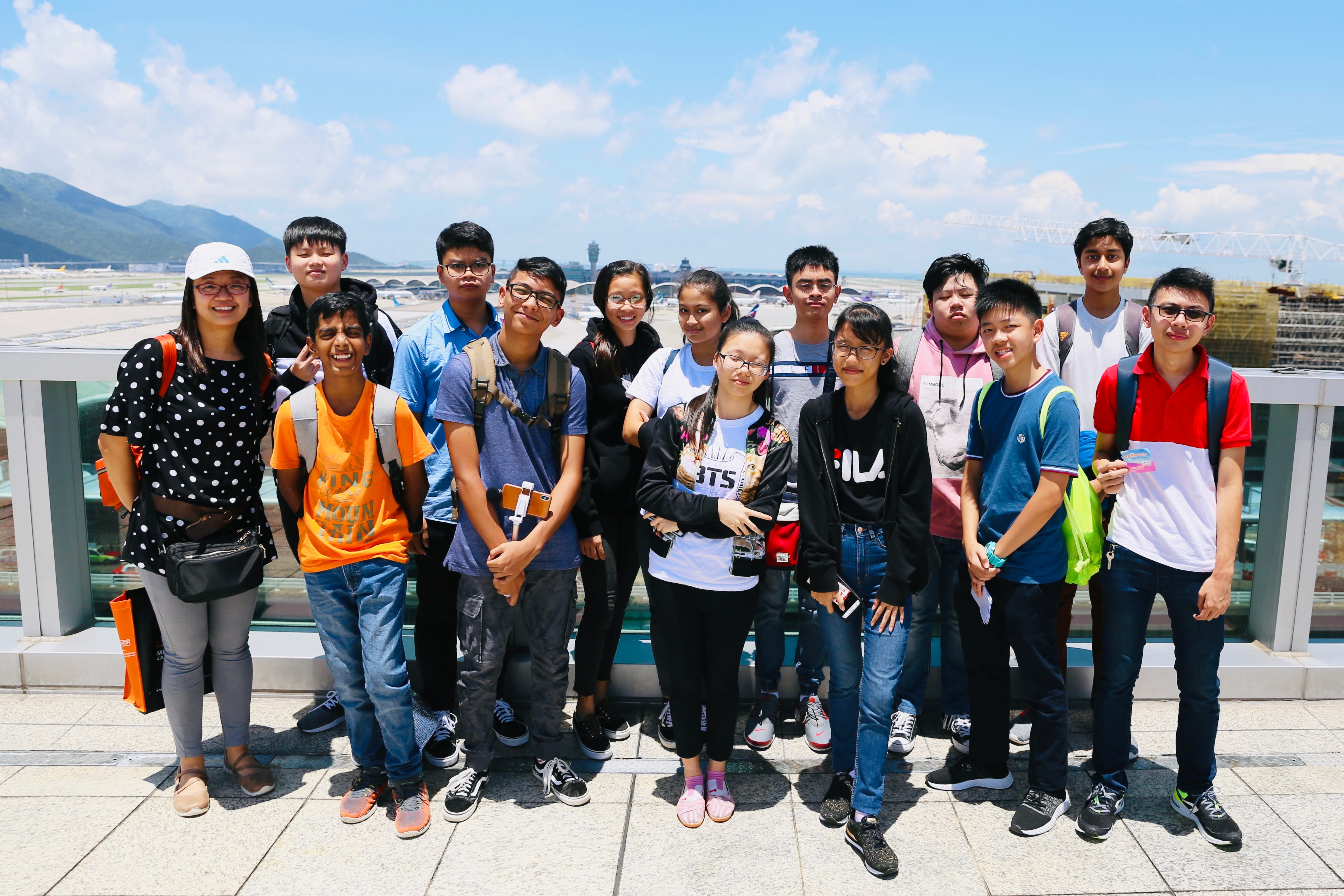 A group of about 15 teenagers posing on an outdoor observation deck overlooking an airport runway on a sunny day.