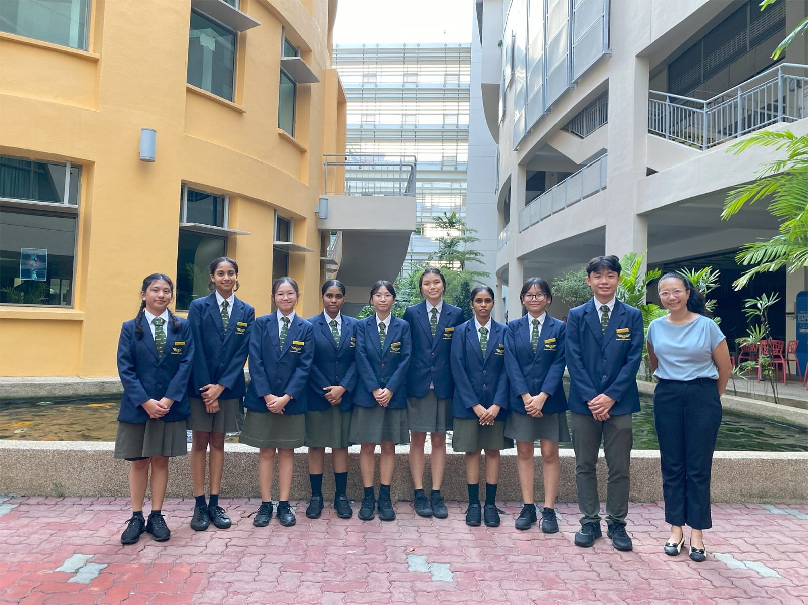 Nine students in navy blazers and green ties stand with a teacher in a school courtyard between multi-story buildings.