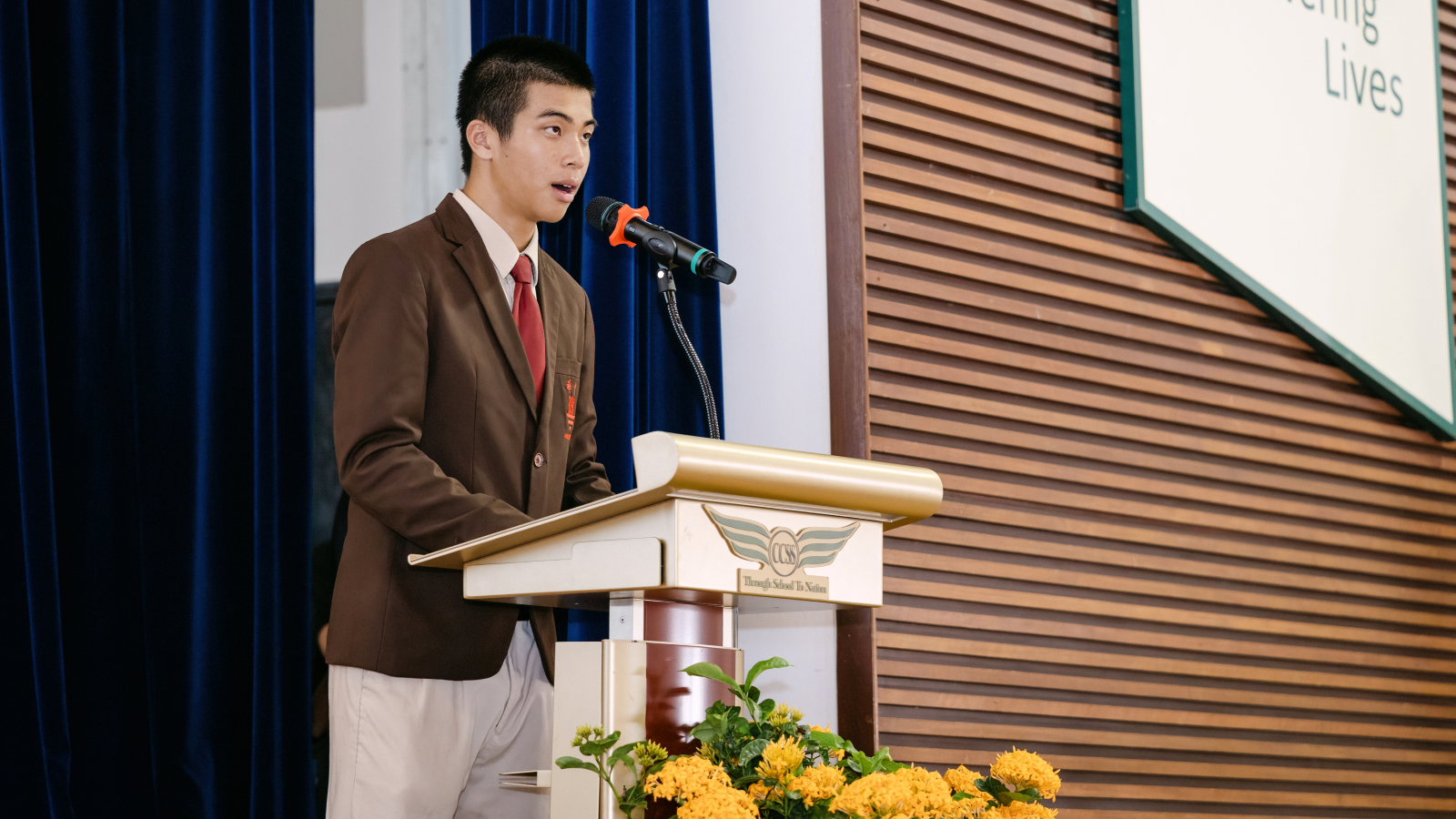 A young male student in a brown blazer and red tie speaks at a podium with a microphone and yellow flowers.