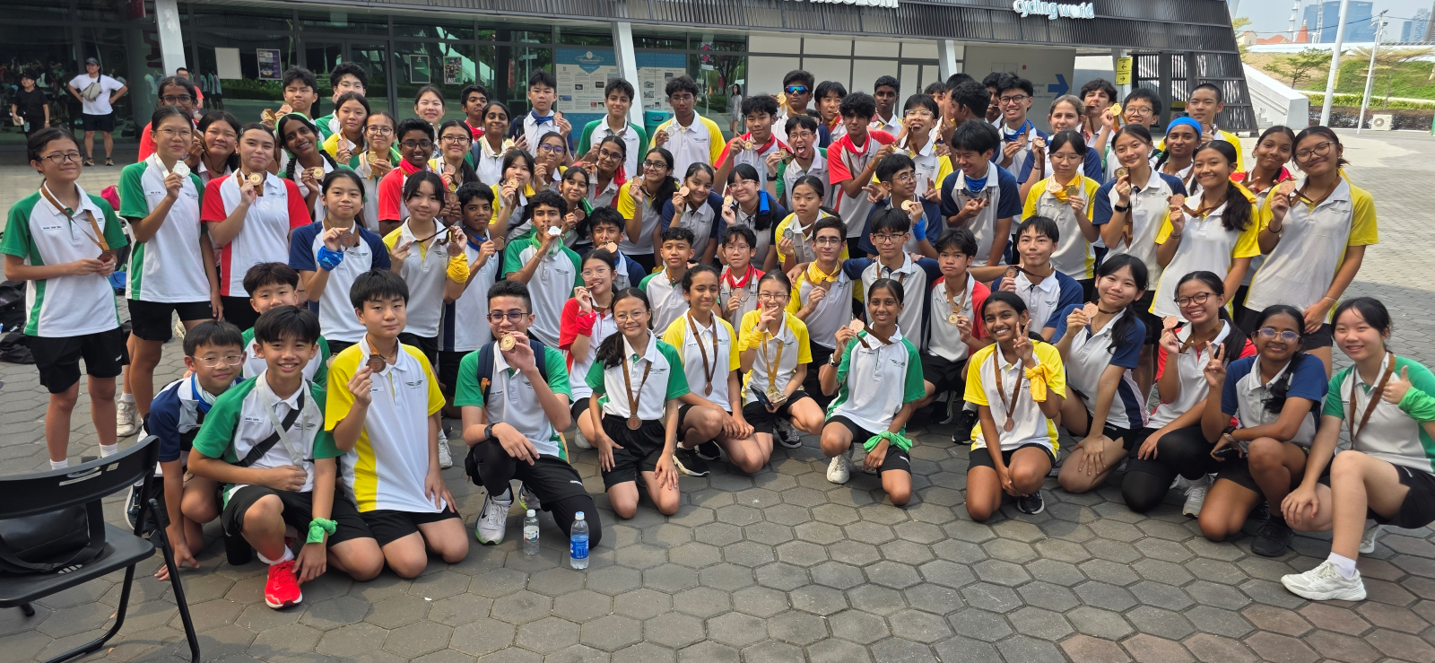 Large group of young students in colorful sports uniforms posing outdoors, many holding up medals and smiling.