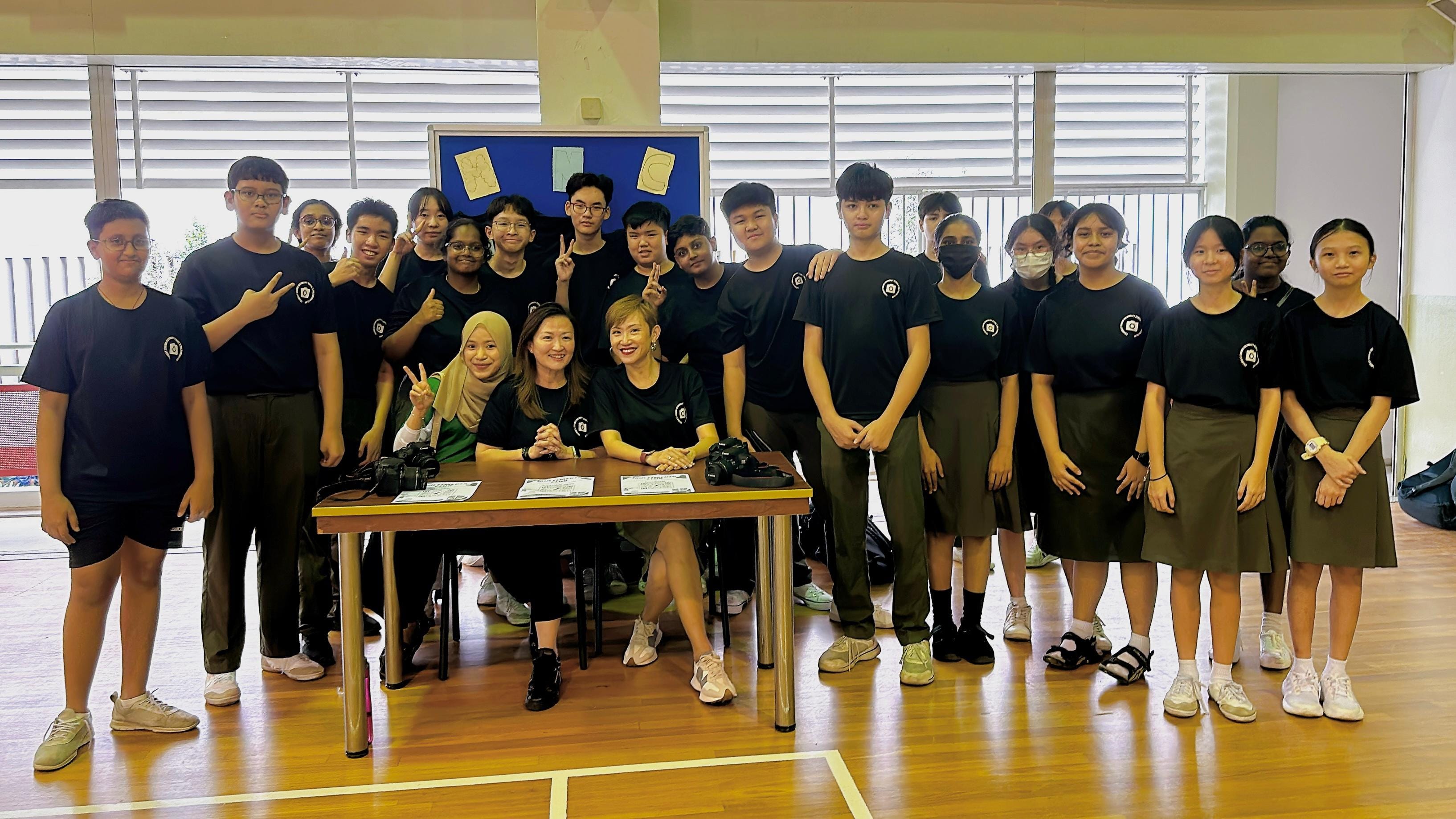 A large group of students in black uniforms pose with two adults seated at a table in a school hall.