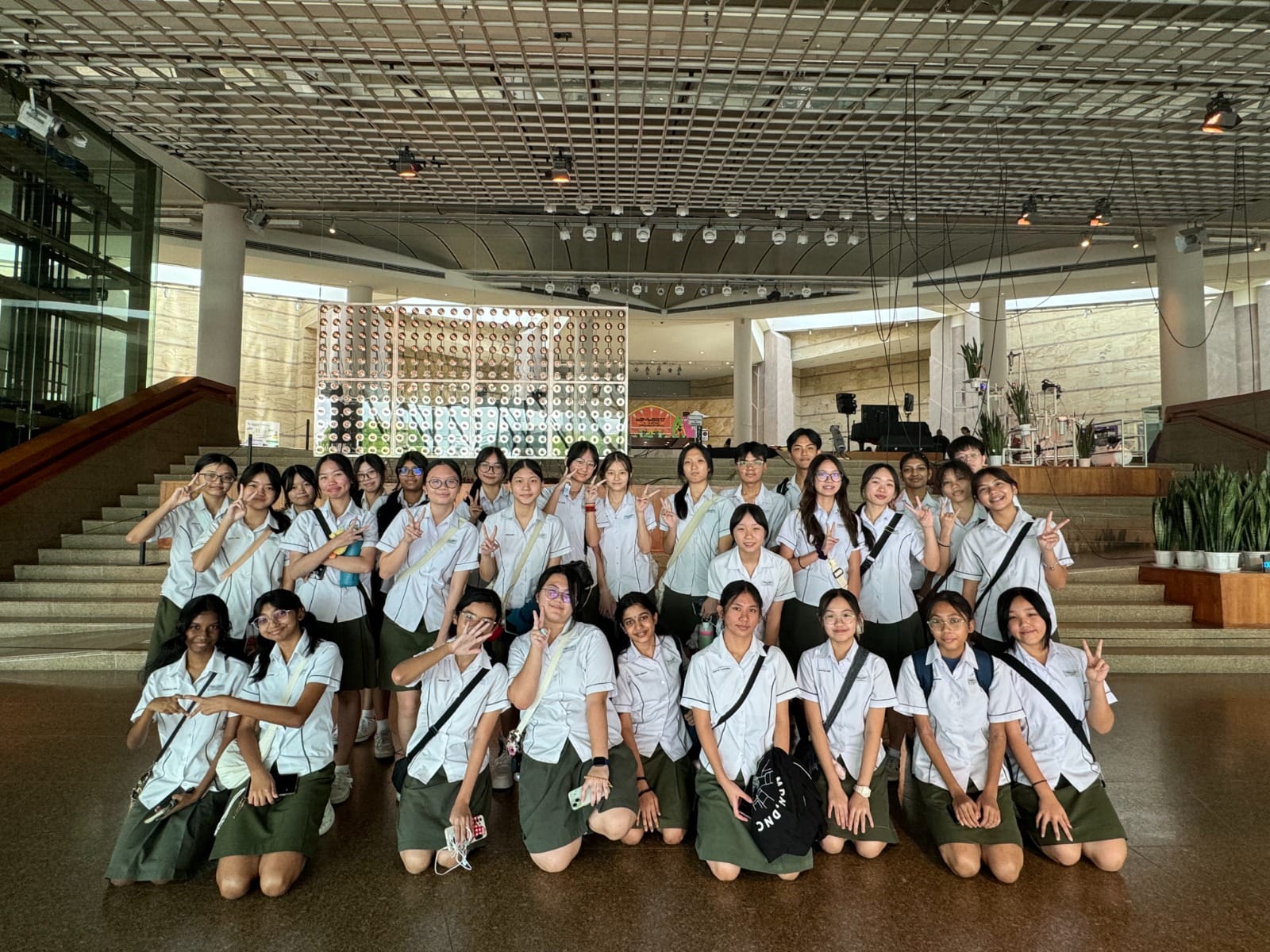 A large group of students in white shirts and green skirts pose with peace signs inside a modern building lobby.