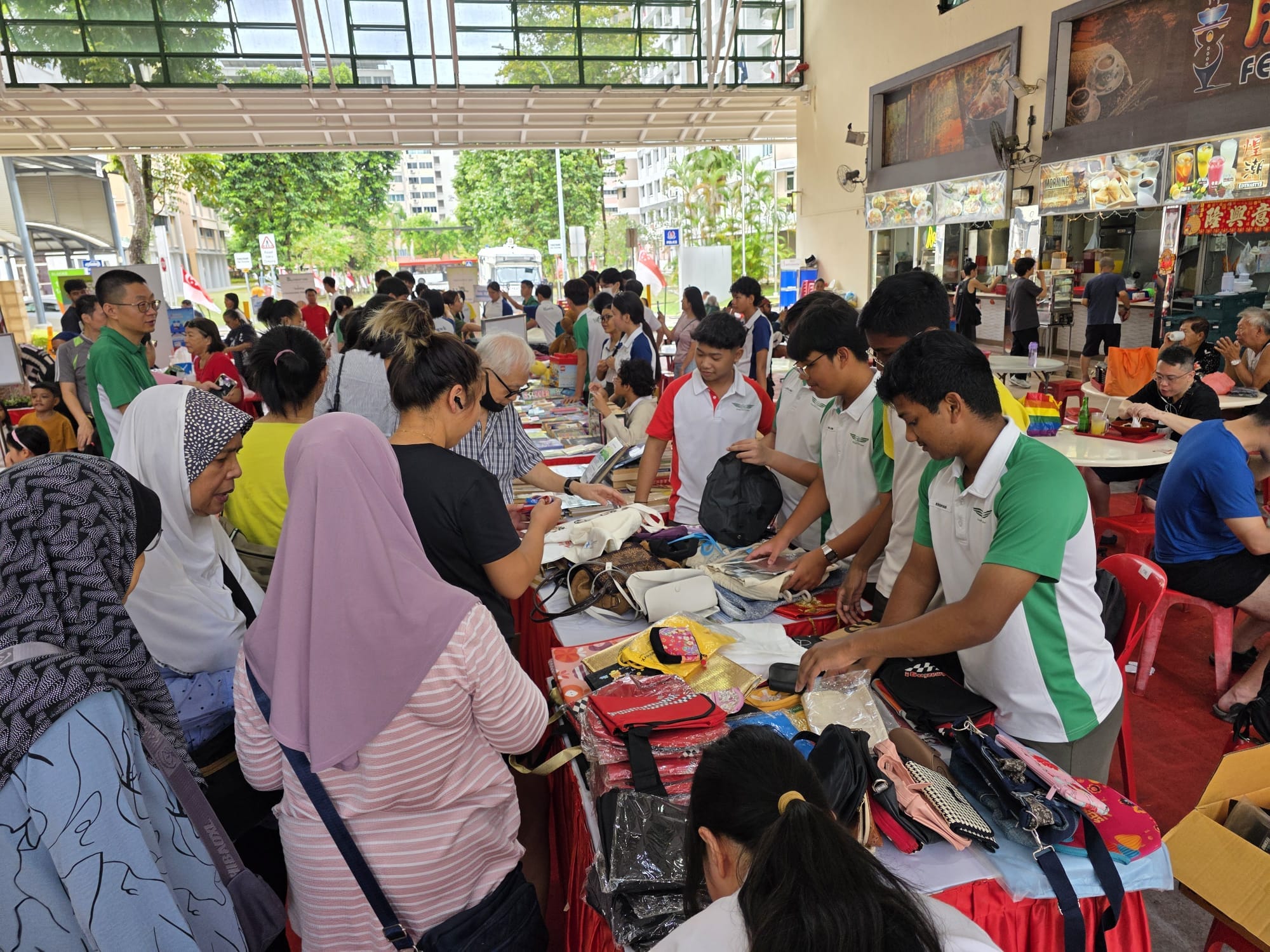 Crowd of people browsing tables filled with bags and accessories at an outdoor community market near shops.