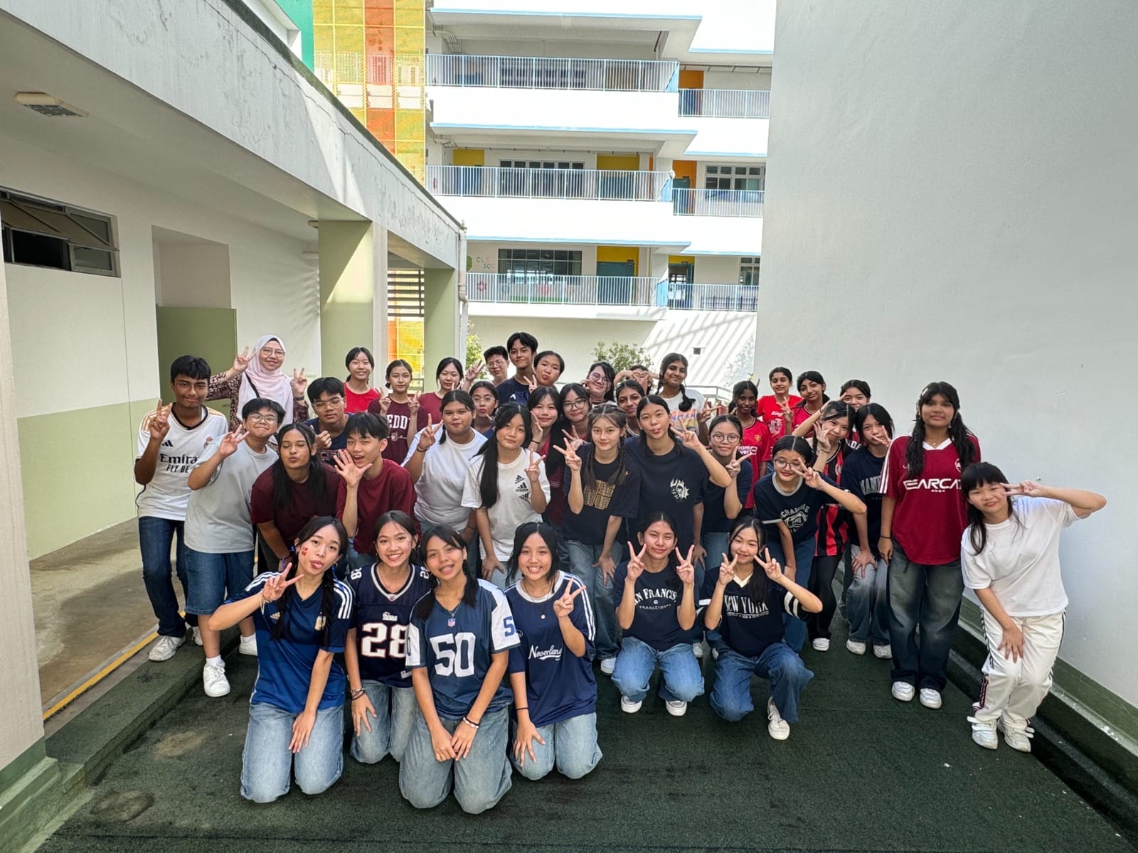 A large group of students posing with peace signs in a school courtyard with multi-story buildings behind them.