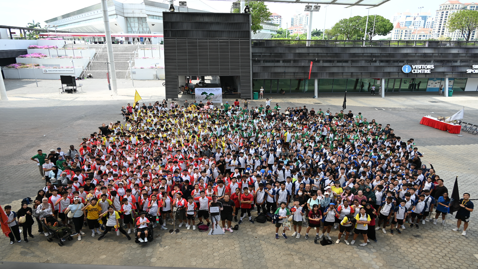 Large group of hundreds of students in colorful uniforms gathered outdoors at a Singapore sports venue for a group photo.