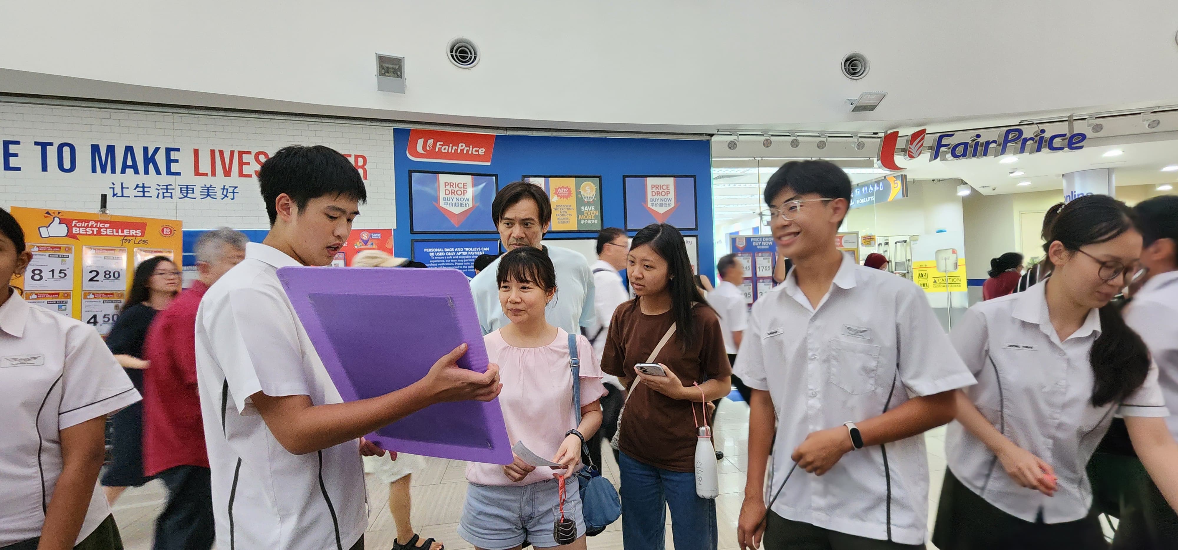 Students in white uniforms interact with shoppers outside a FairPrice supermarket, one holding a purple clipboard.