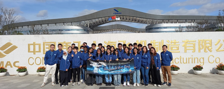 A group of students in blue jackets pose with a banner in front of the COMAC manufacturing facility in China.