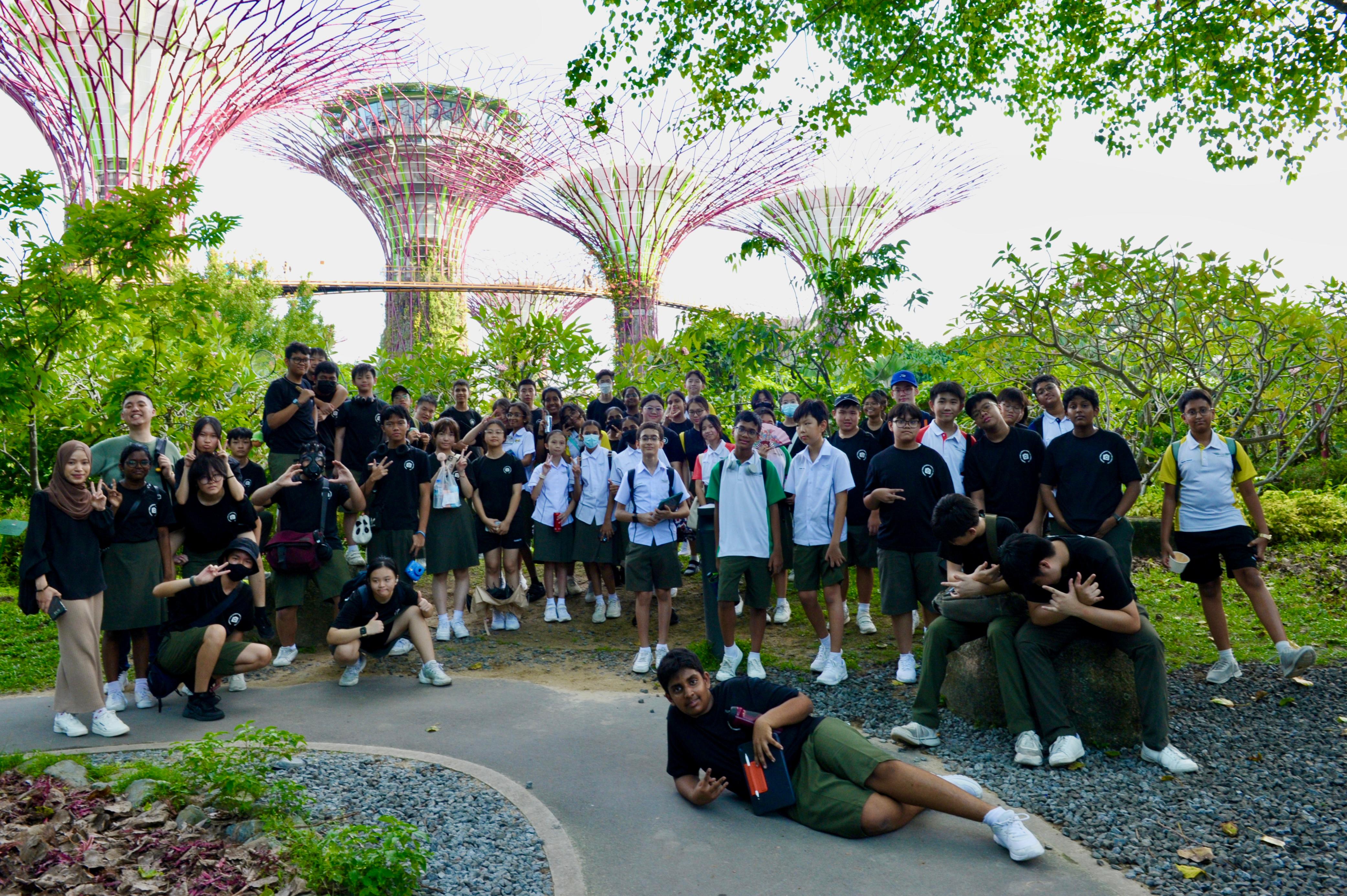 A large group of students posing outdoors at Gardens by the Bay, Singapore, with towering Supertree structures behind them.