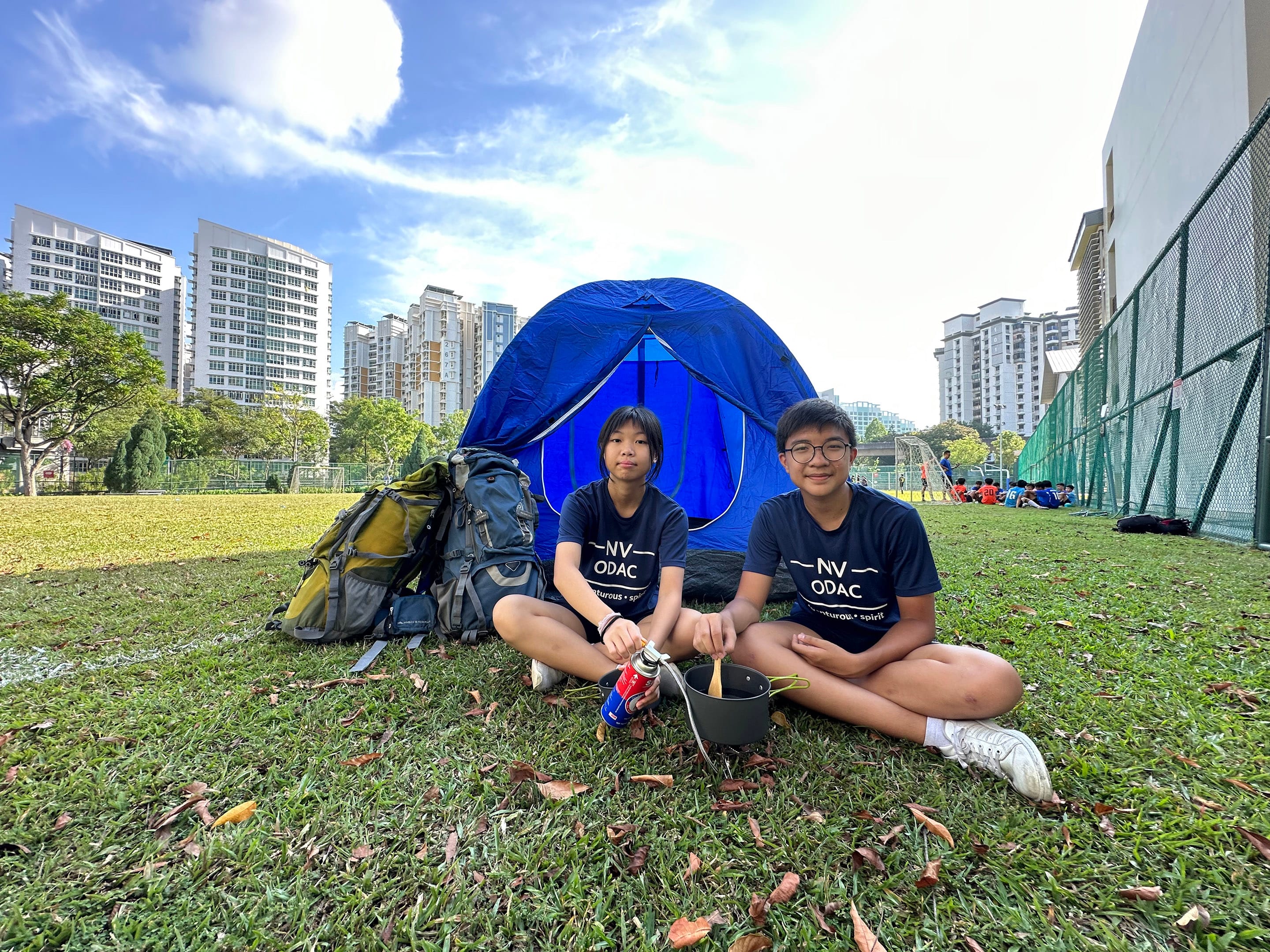 Two NV ODAC students sit on grass cooking beside a blue tent and backpack, on school field.