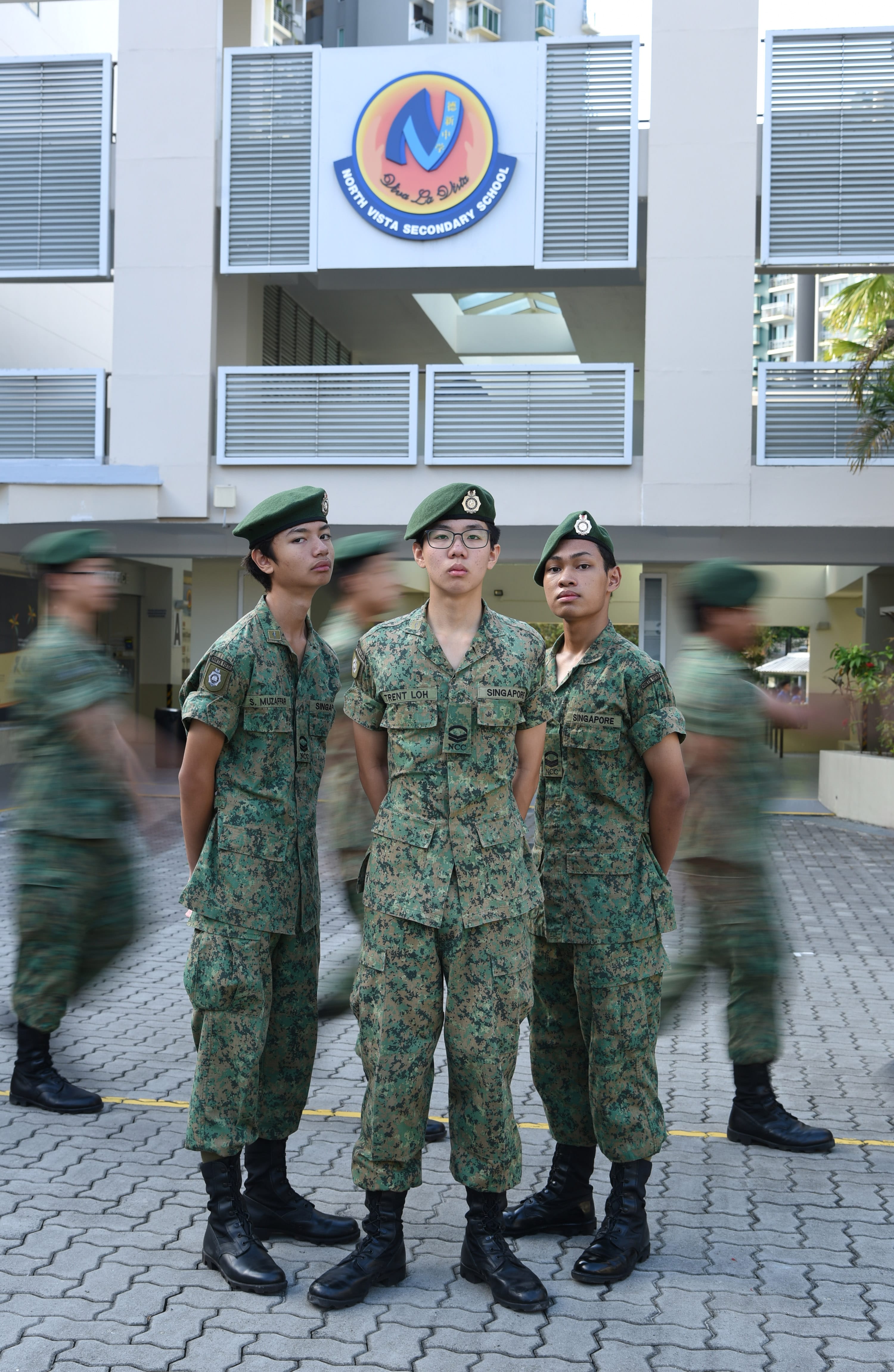 NV NCC cadets in uniforms and green berets stand in front of North Vista Secondary School logo