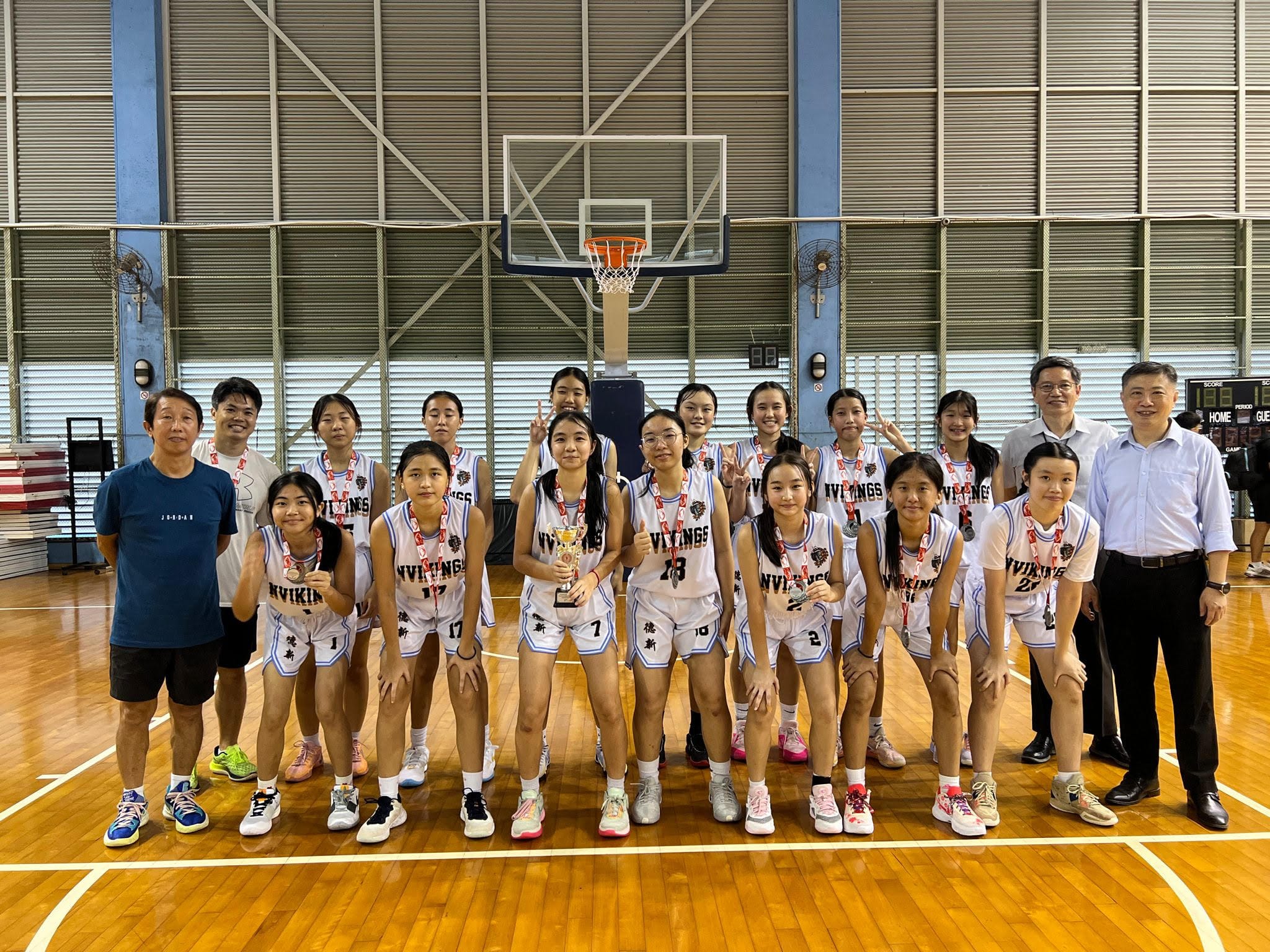 NV Girls basketball team in white jerseys posing with medals and a trophy on an indoor court under a hoop.