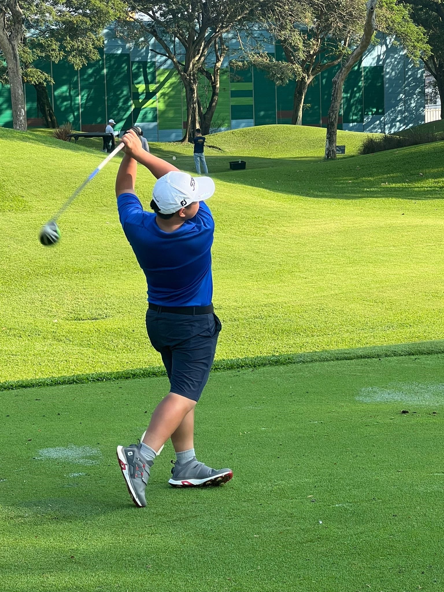 NV golfer in a blue shirt and white cap completes a full swing on a green golf course.