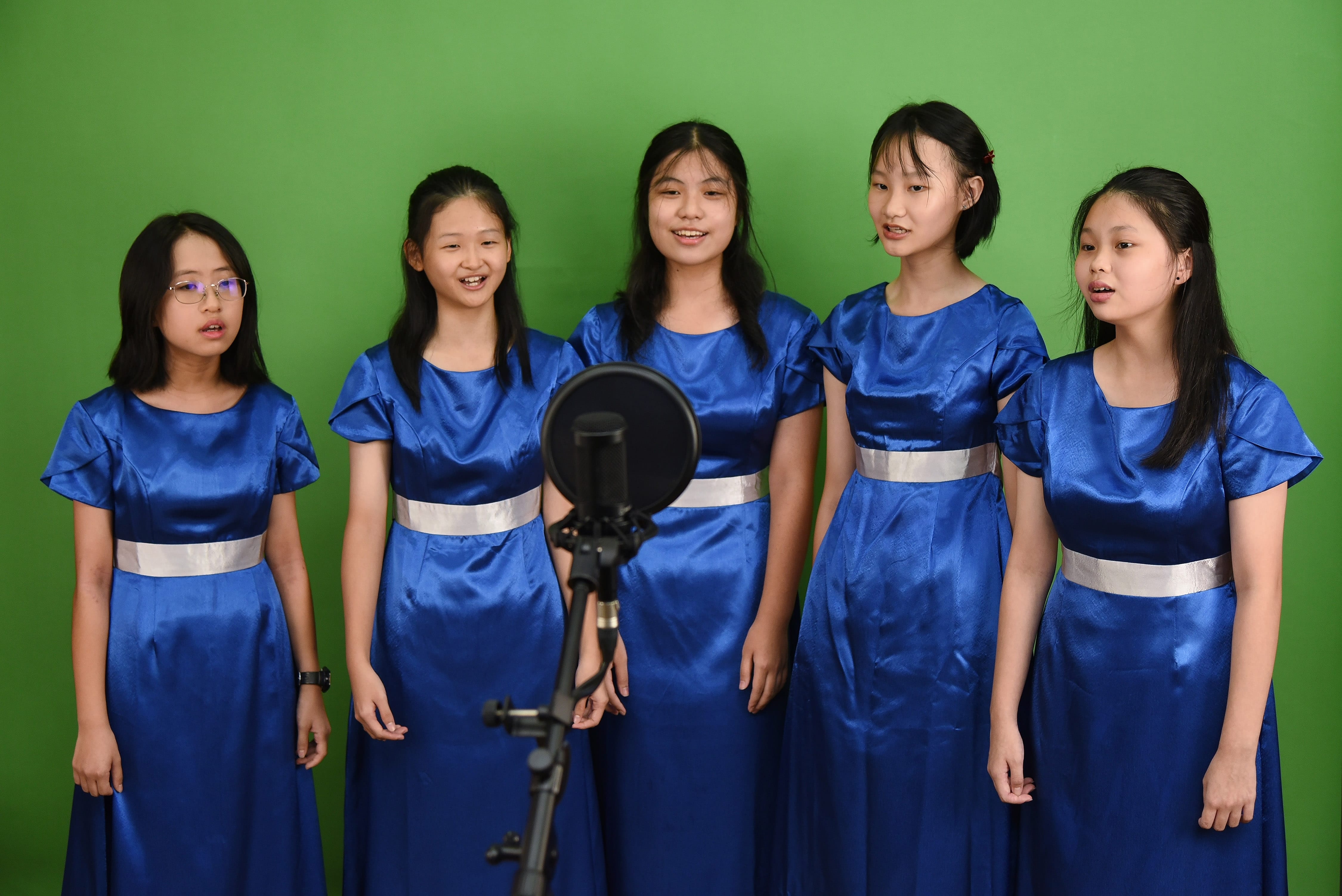 NV Choir members sing into a microphone on a stand before a green background.