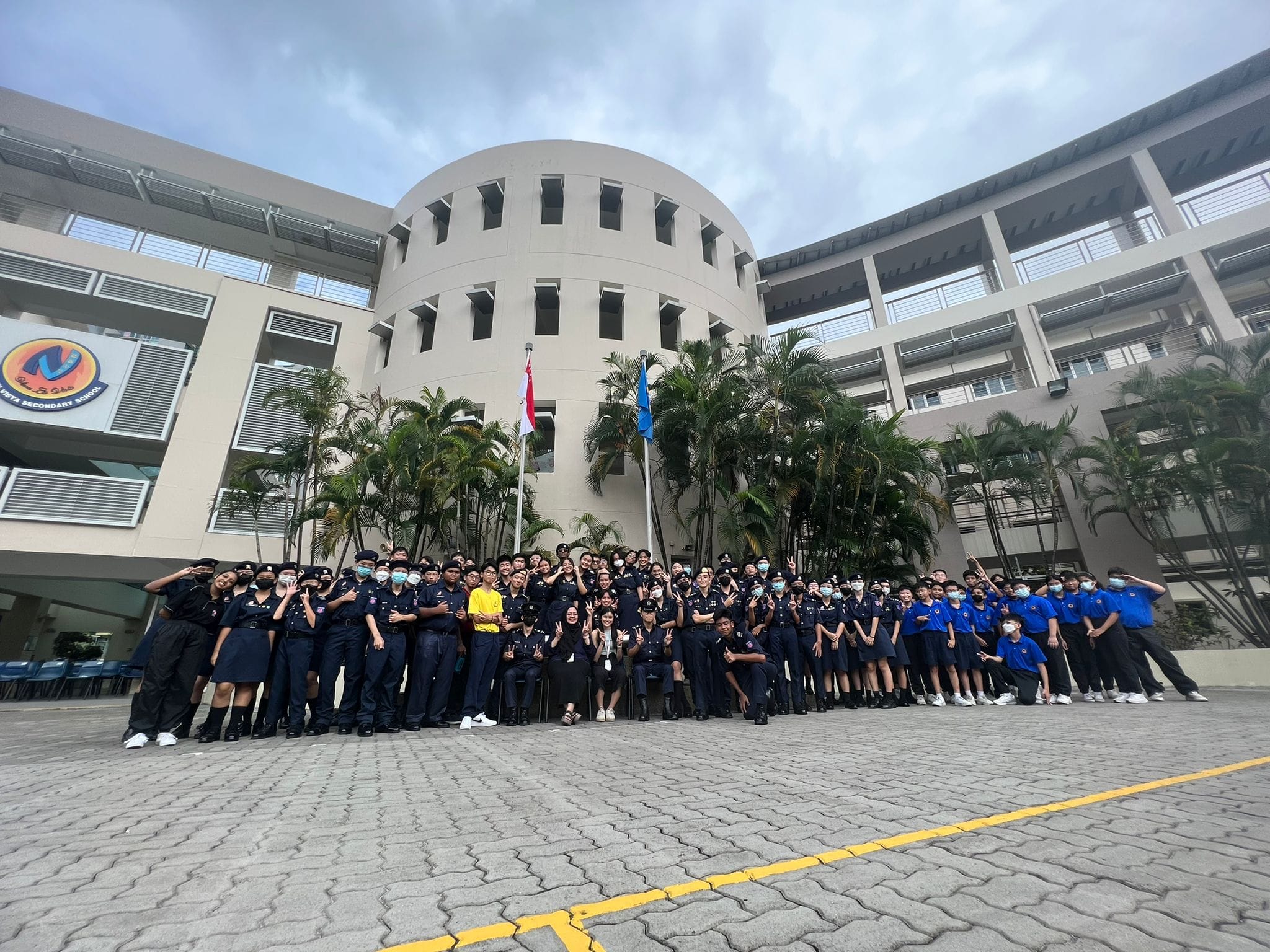 NV NPCC cadets in uniforms posing outdoors in front of the flag poles in the school parade square.