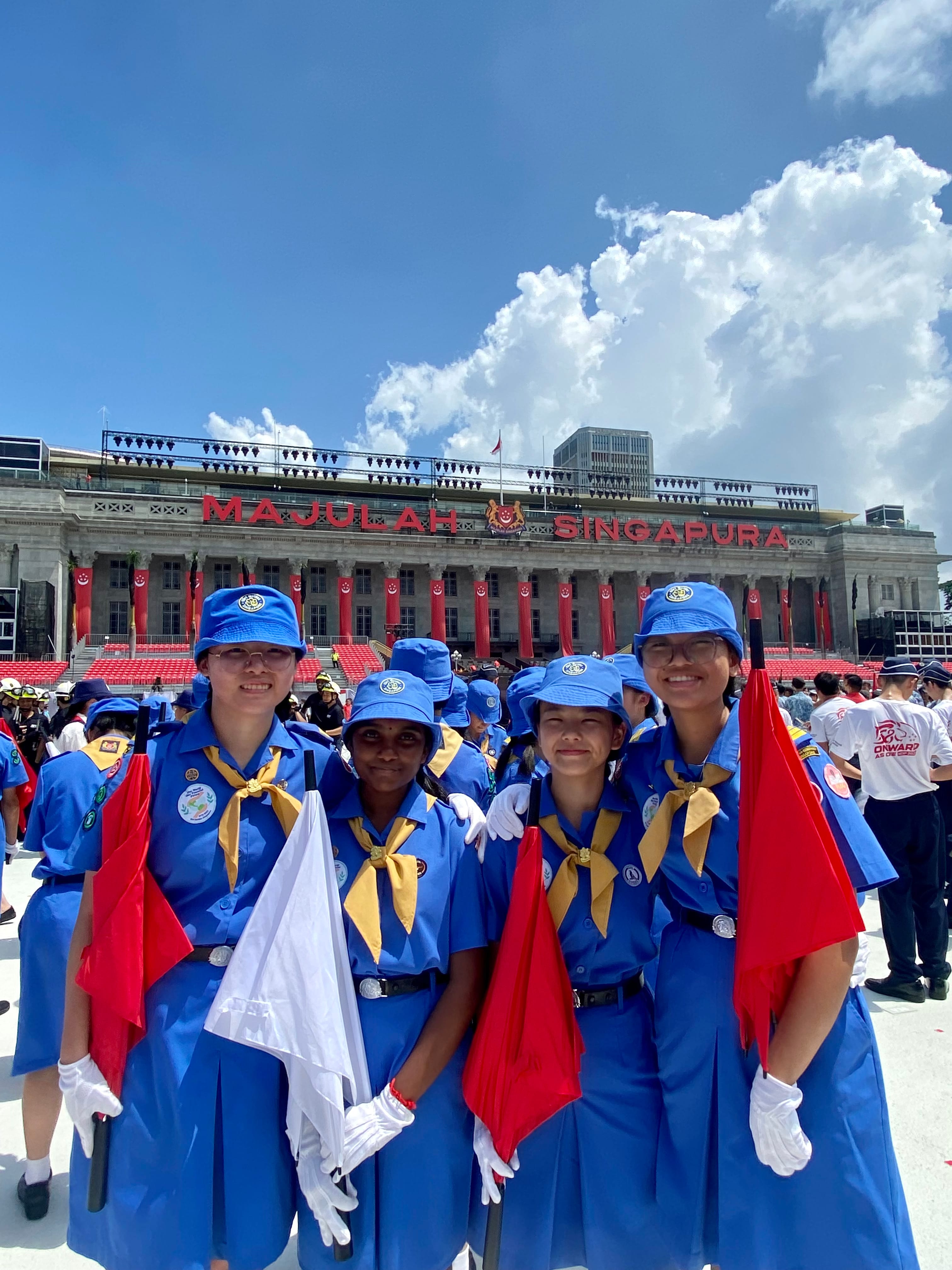 NV Girl Guides members hold flags in front of a building bearing the words 'Majulah Singapura'.