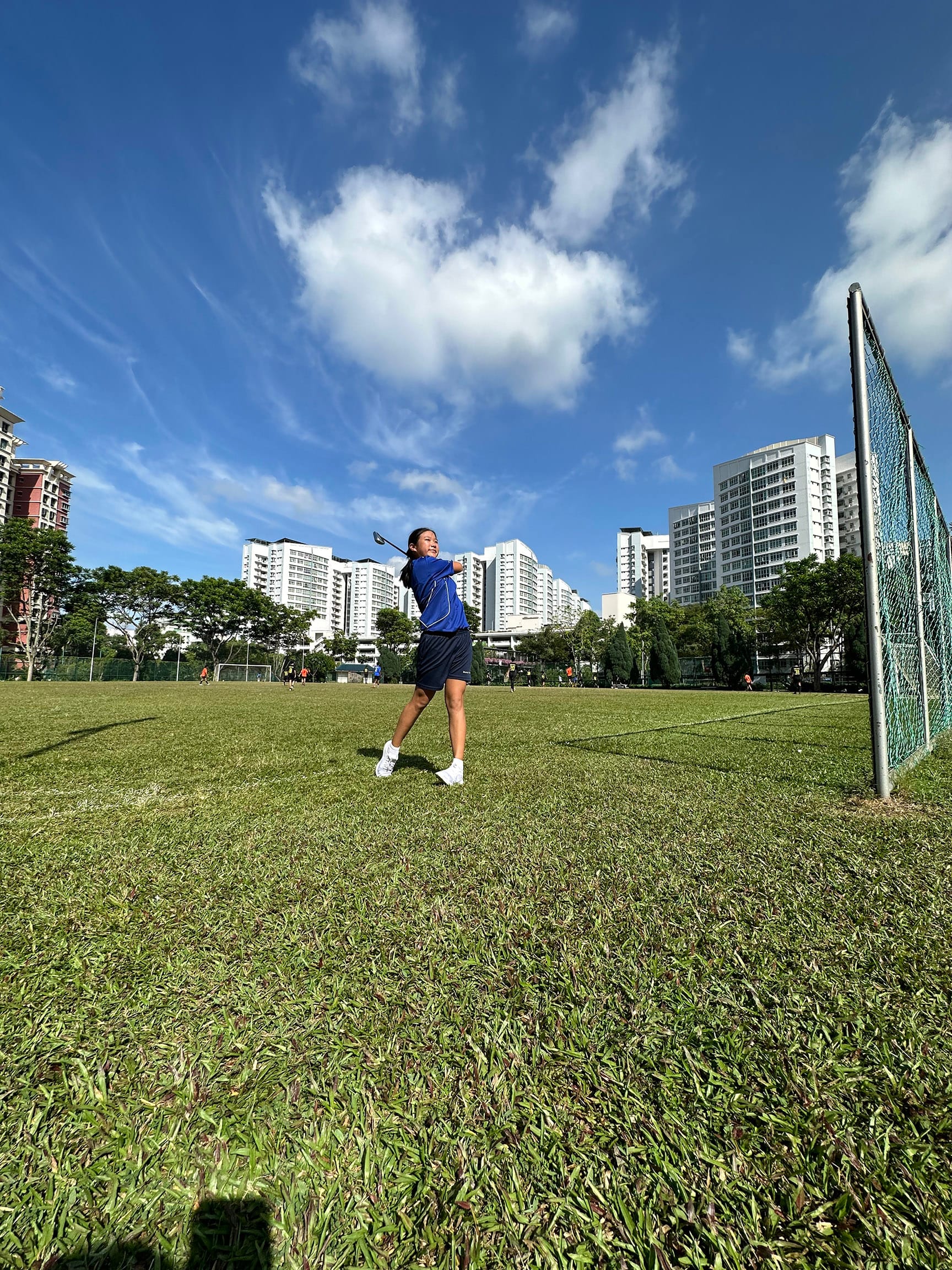NV golfer in a blue shirt swings a golf club on a green field with tall white buildings under a blue sky.