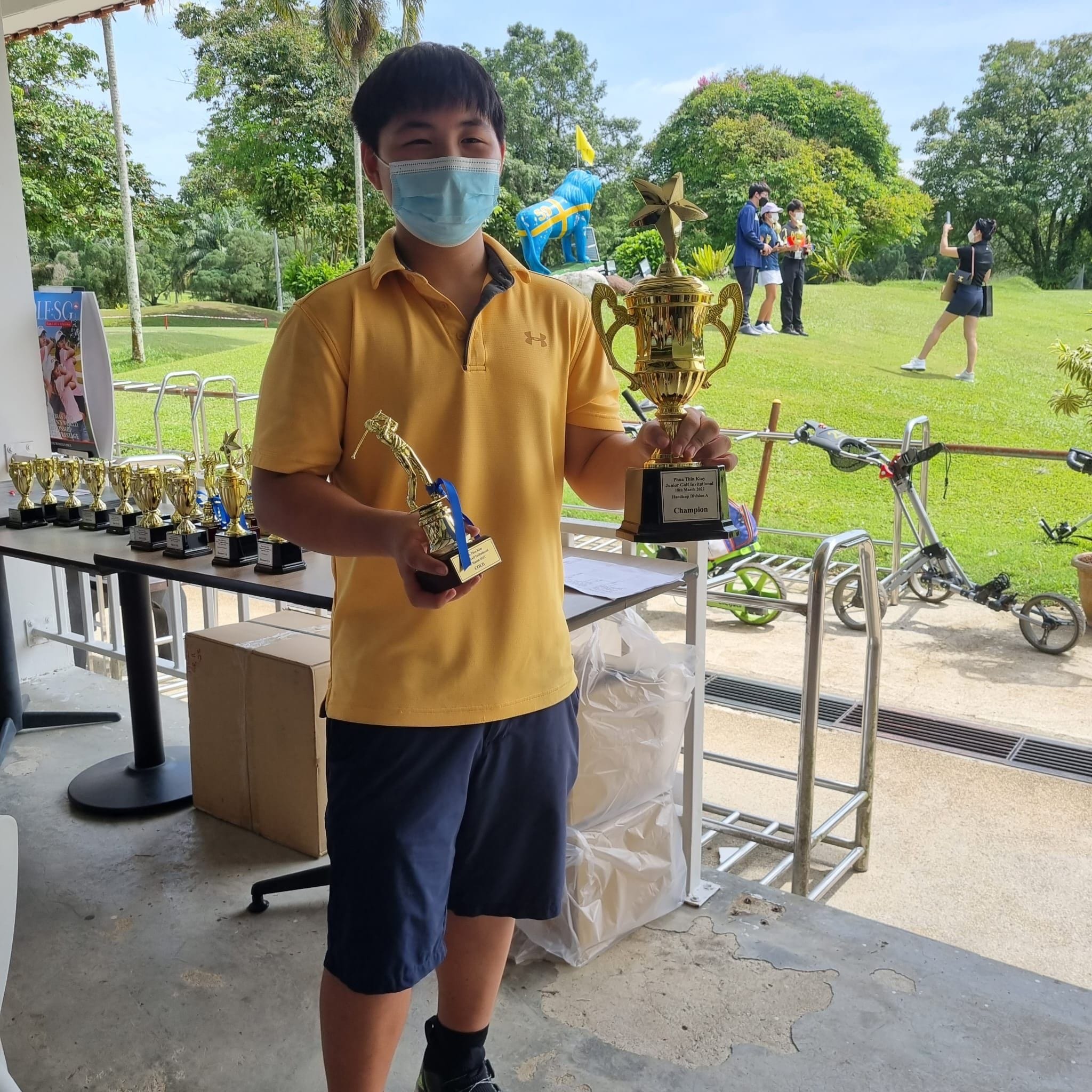 NV golfer holding two gold trophies at a golf course with more trophies on a table behind him.