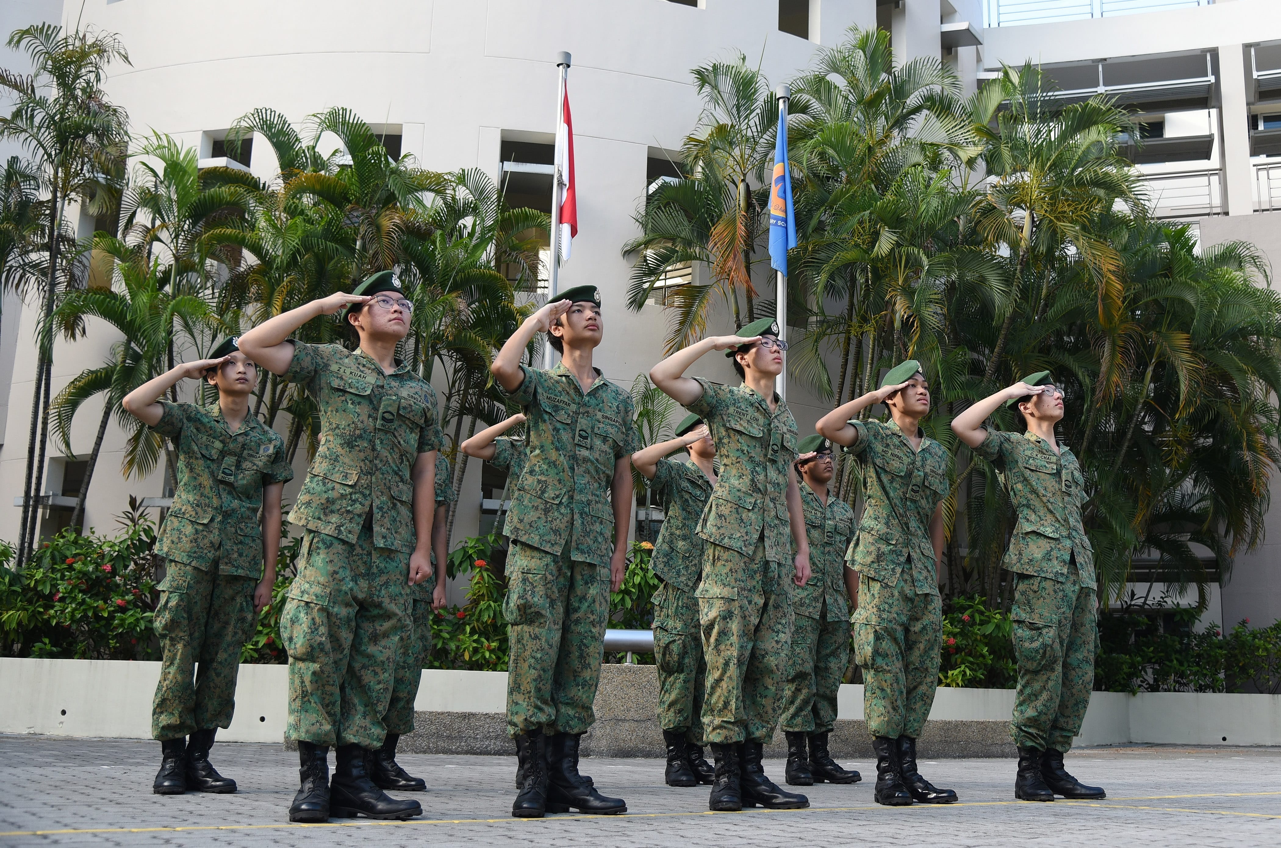 NV NCC cadets in camouflage uniforms and green berets saluting near a flagpole at the parade square..