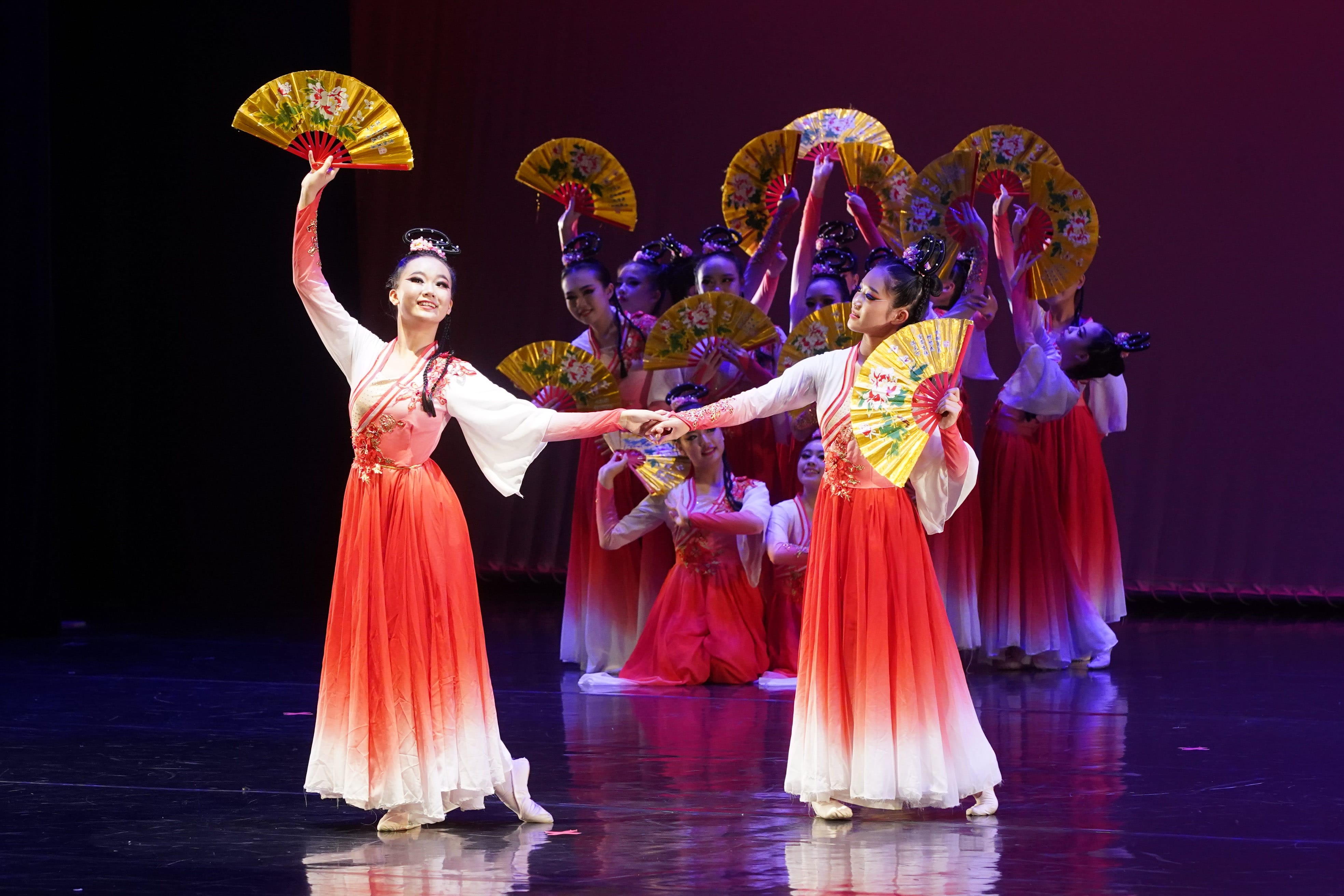 A group dancers in red and white traditional Chinese costumes perform on stage holding gold decorative fans.