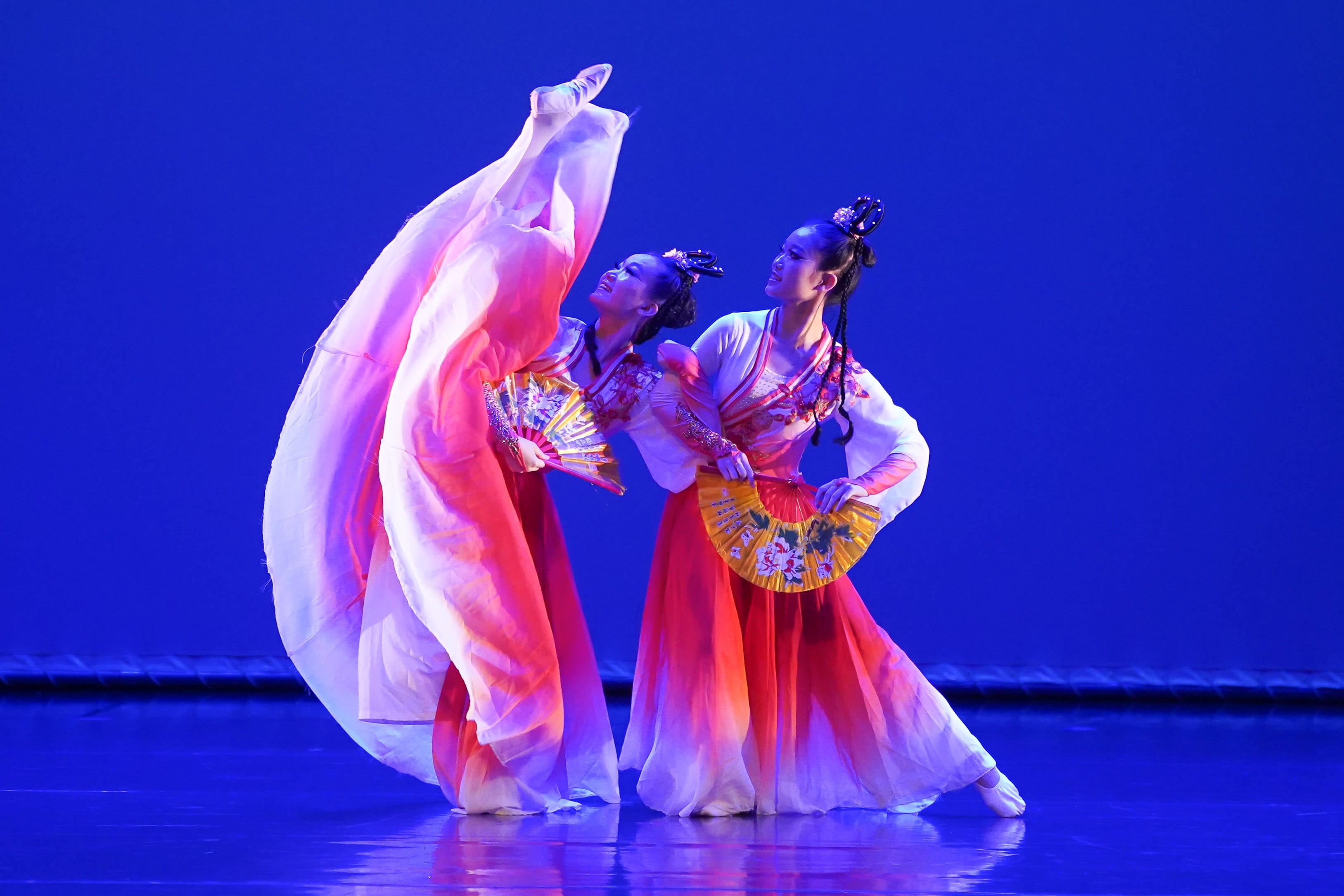 Dancers in red and white traditional Chinese costumes perform with decorative fans on a blue-lit stage.