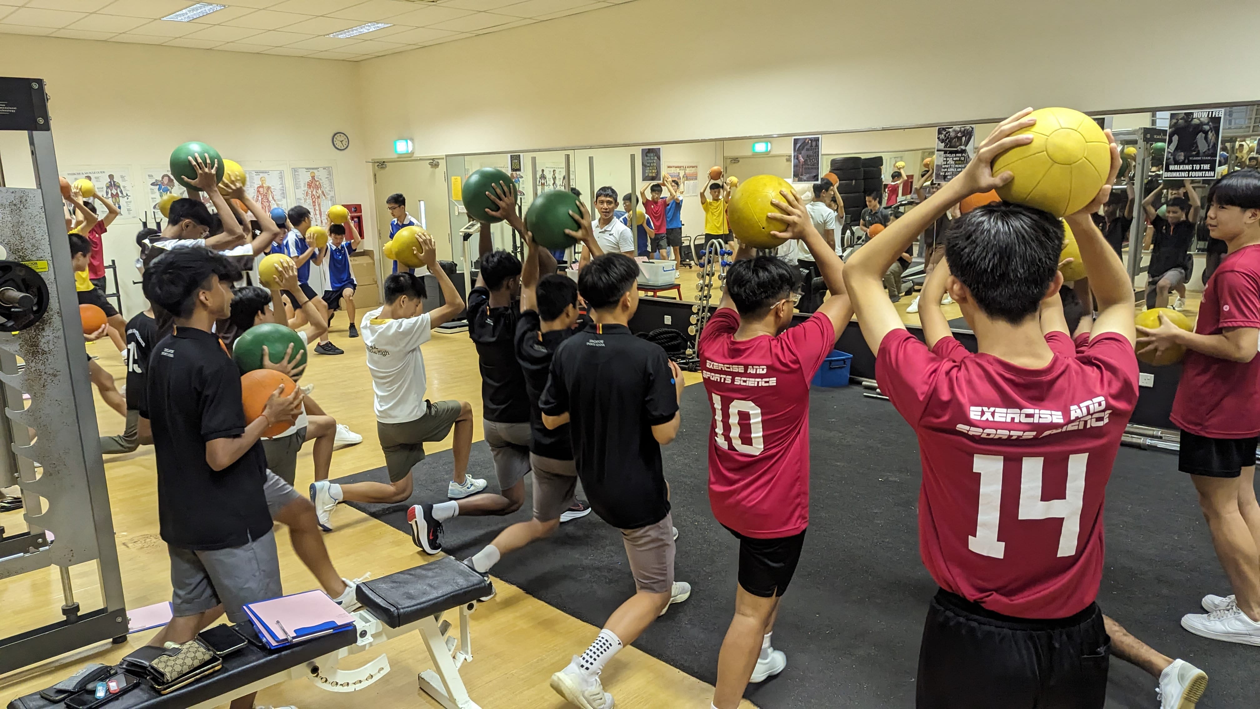 NV students in a gym do lunges while holding medicine balls overhead, wearing Exercise and Sports Science jerseys.