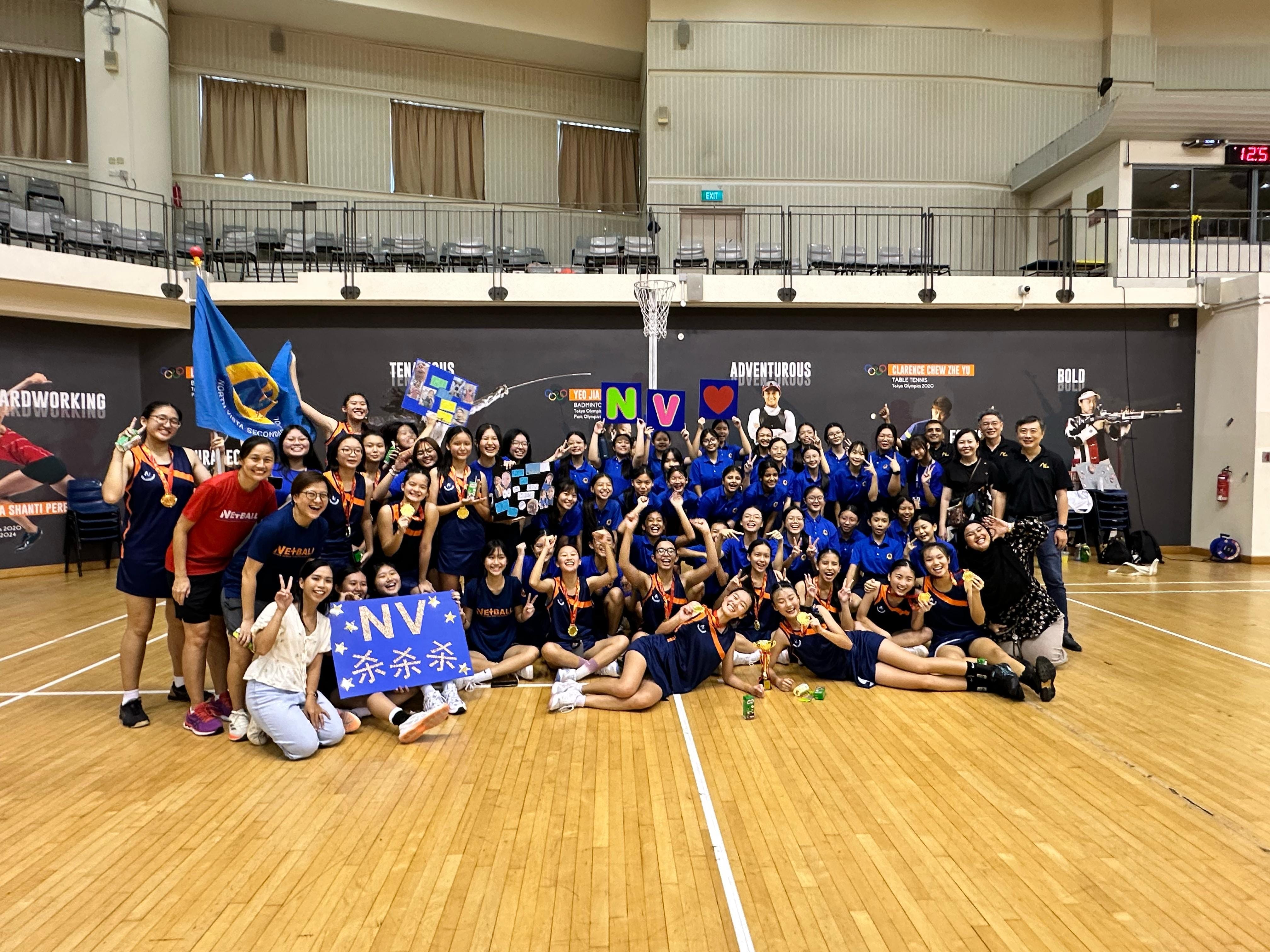 NV netball girls in blue netball uniforms celebrating on a court, holding signs and NV flag.