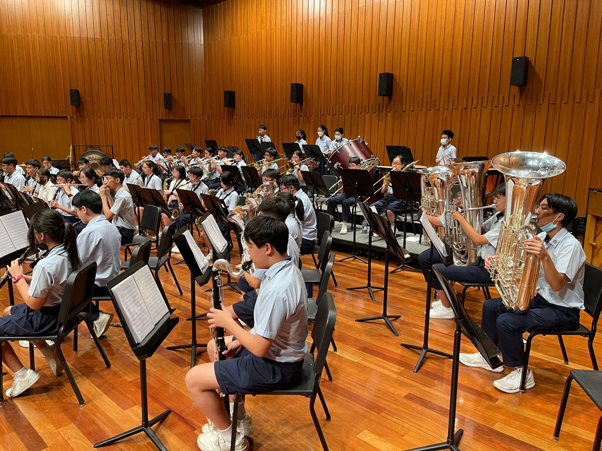 Bnd members rehearse  in a wood-paneled hall, playing tubas, clarinets, and brass instruments.