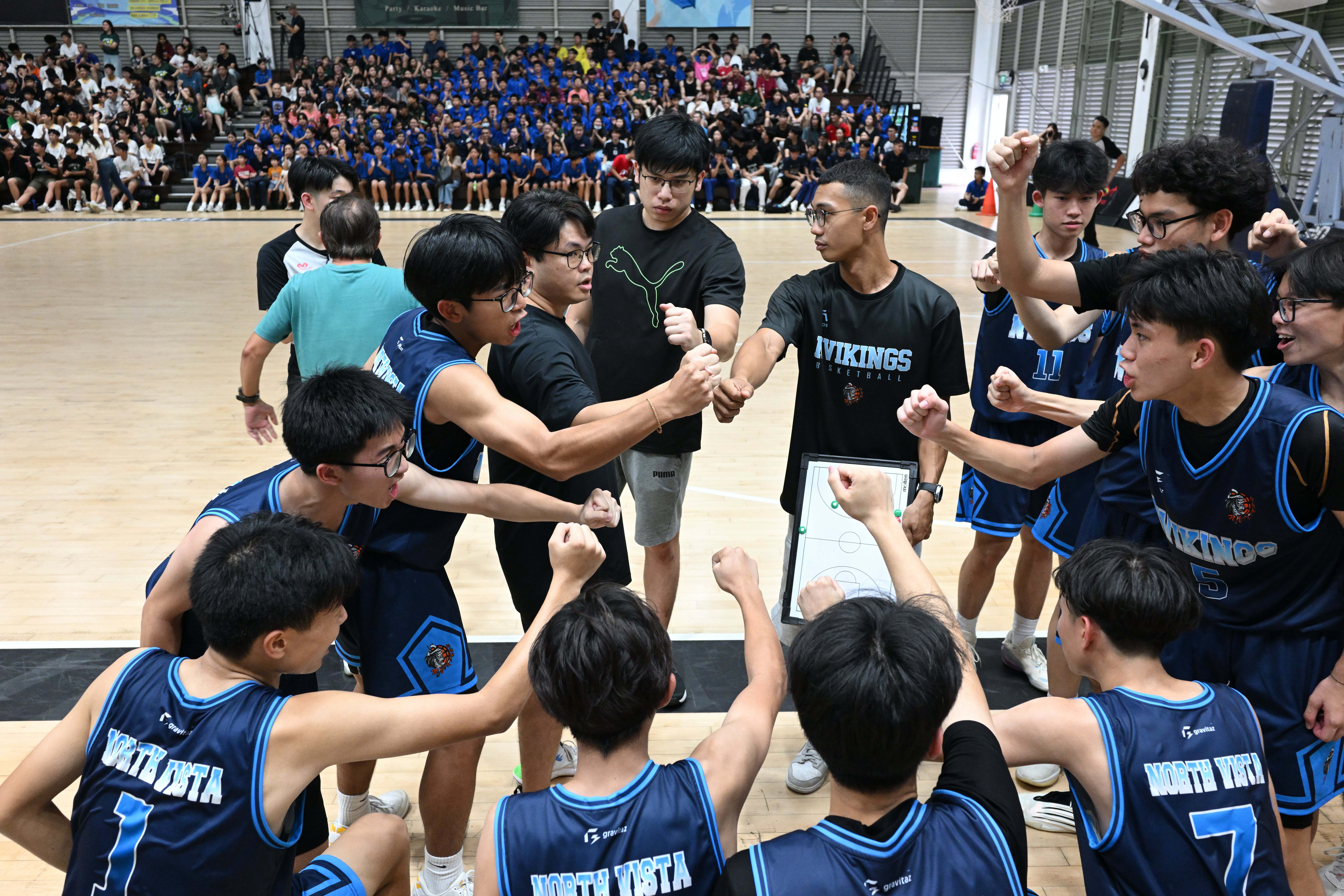 North Vista basketball players and coaches huddling up with raised fists on the court, with a packed crowd watching.