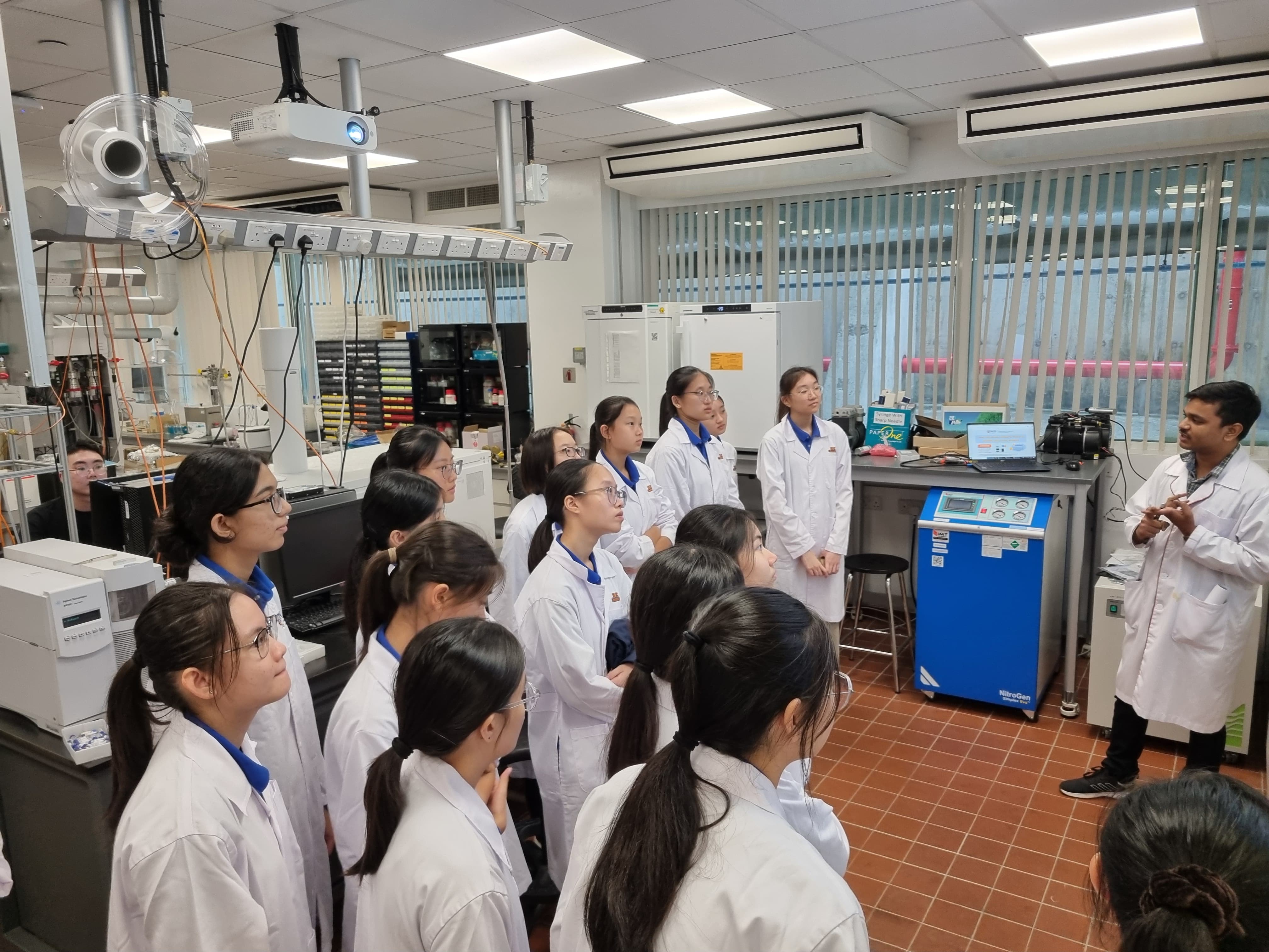 A man in a white lab coat speaks to NV students in white coats inside a science laboratory.