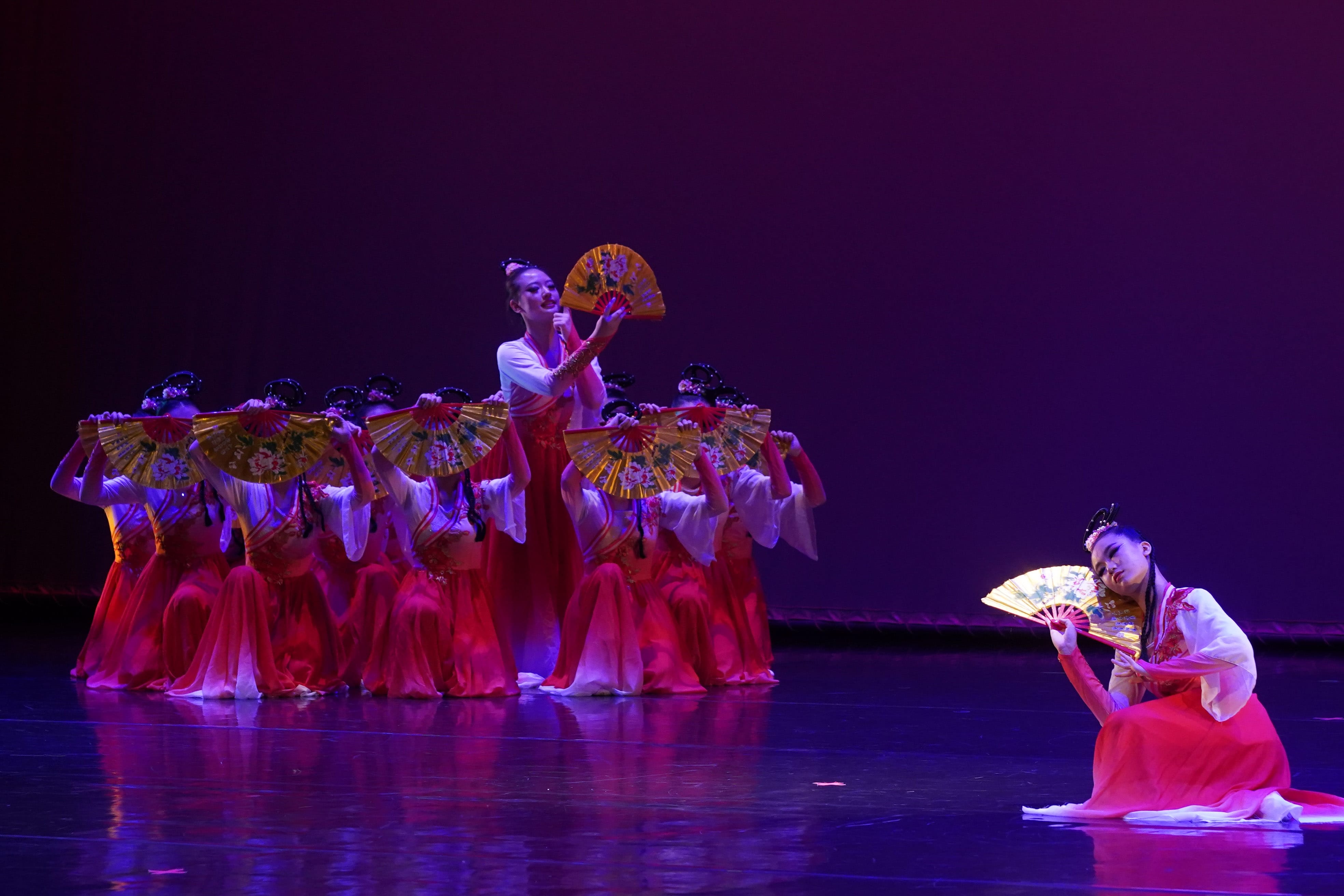 Dancers in red and white traditional Chinese costumes perform a fan dance on a purple-lit stage.