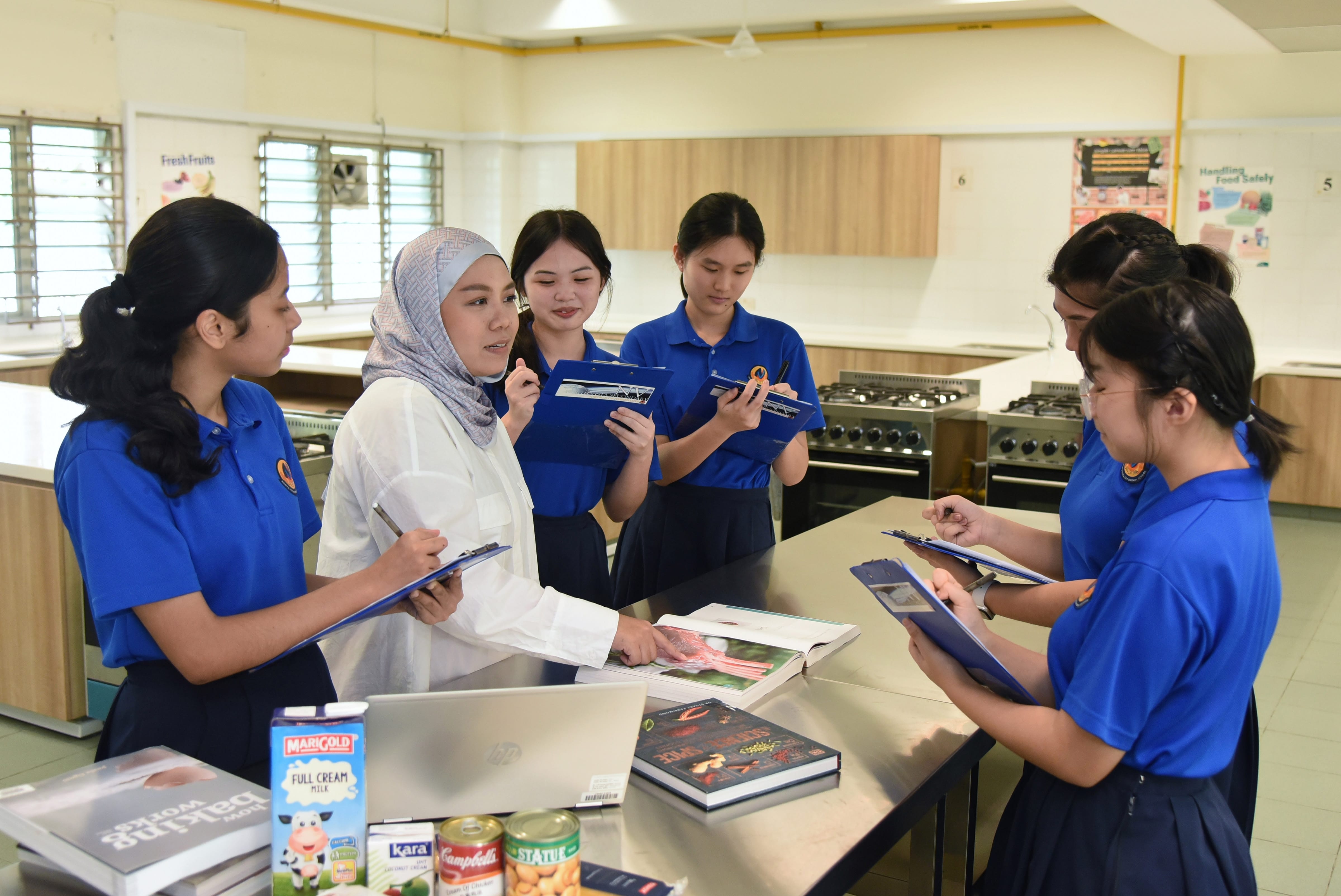 NV students gather around a kitchen classroom counter with clipboards, books, and food items listening to the teacher explaining