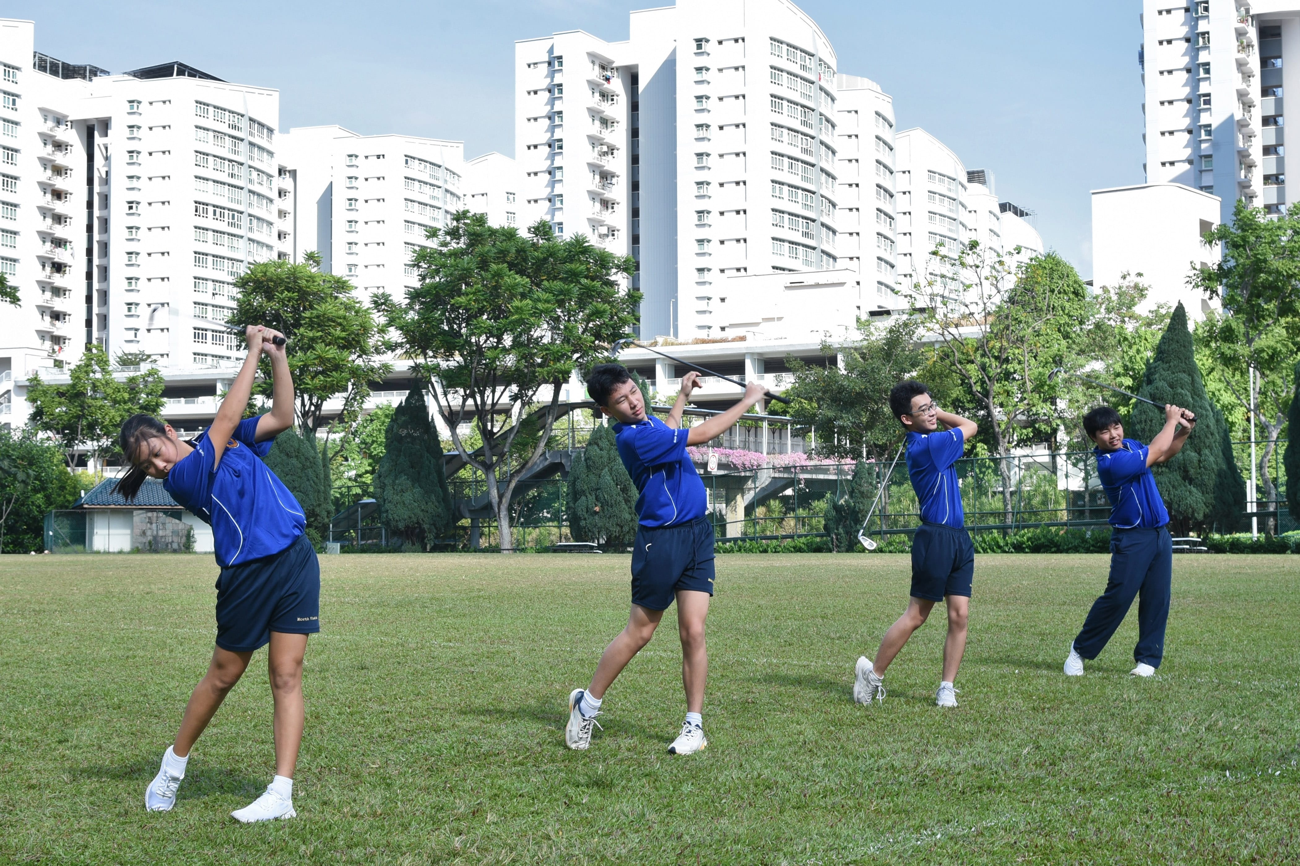 NV golfers in blue uniforms practice golf swings on a grass field with tall white buildings behind them.