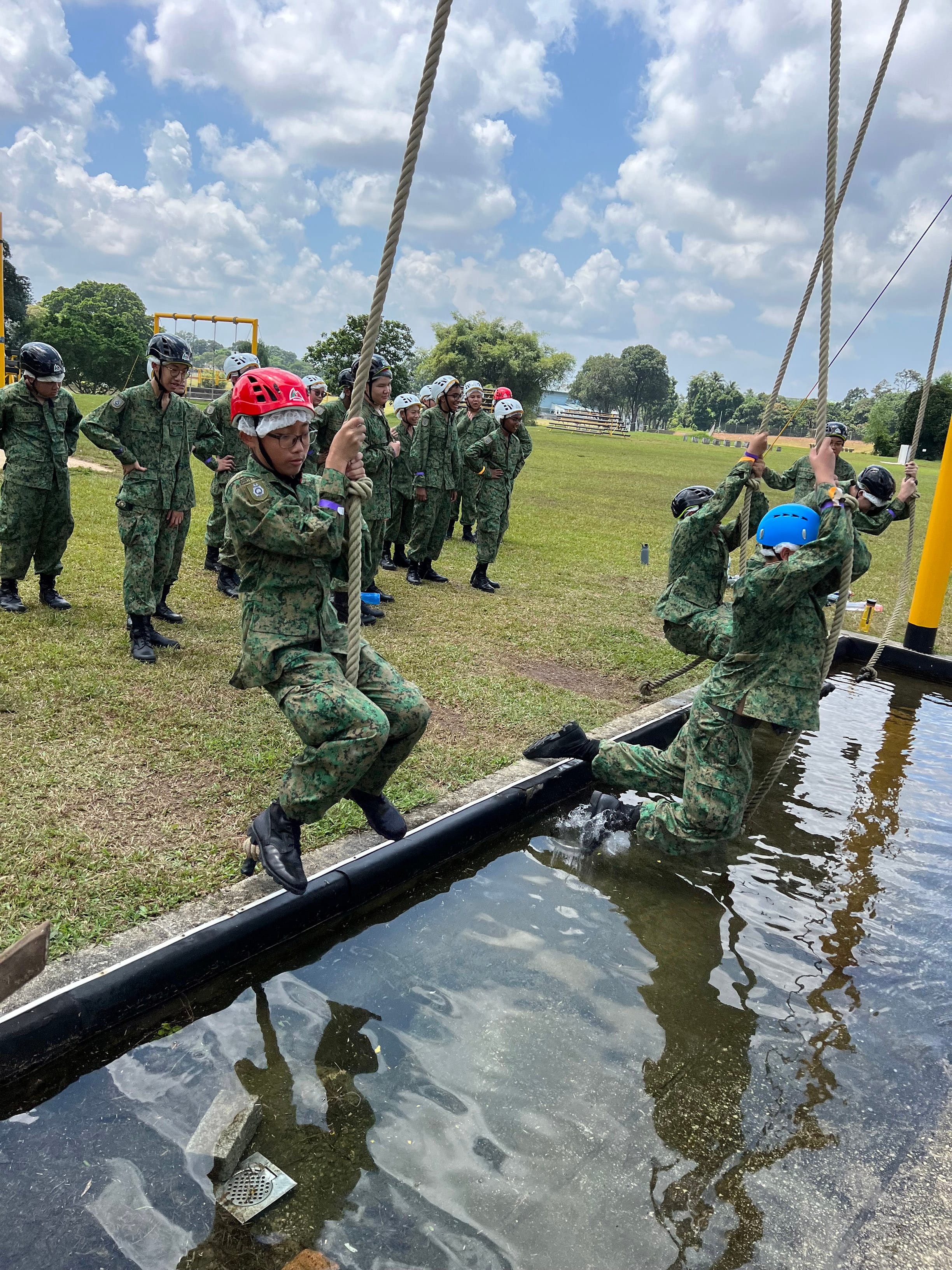 NV NCC cadets cross a water obstacle using ropes while others waiting for their turn on a grassy field.