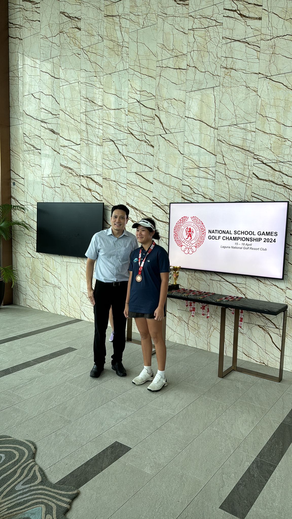 NV student golfer wears a medal pose beside a screen showing National School Games Golf Championship 2024.